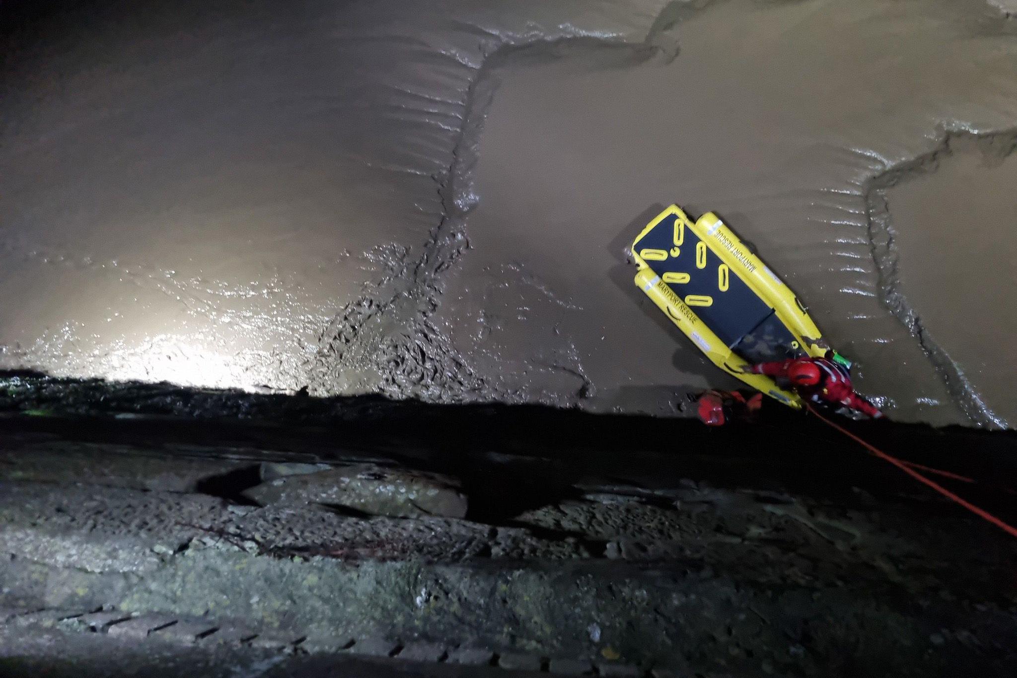 Two rescuers on fluorescent yellow inflatable lifeboat are pulled up at a bottom of a grey stone wall. The scene is lit by torch. 