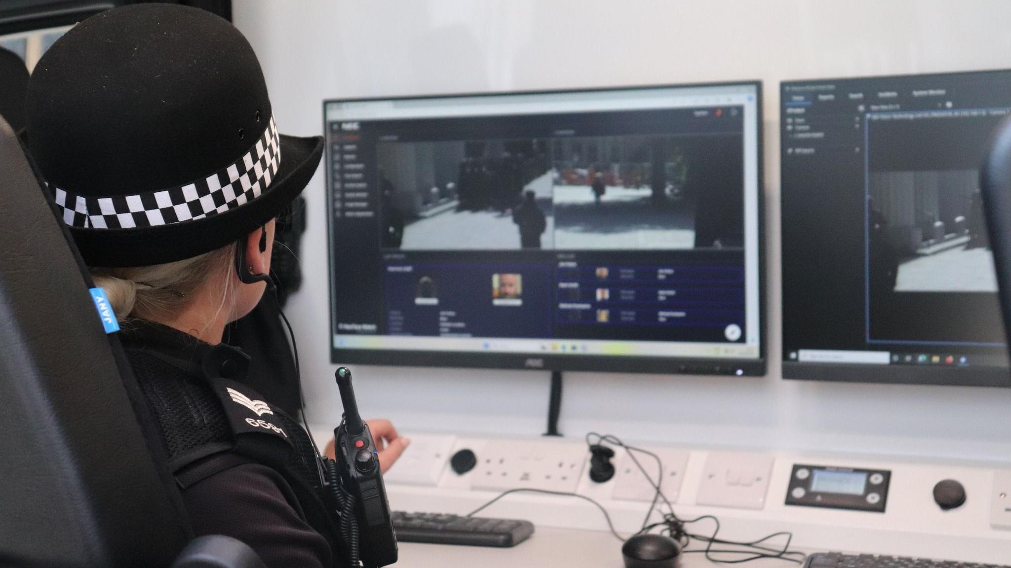 A female PC, in full police uniform, with a walkie talkie, looking at two screens, with a keyboard and mouse. She is turned away from the camera. 