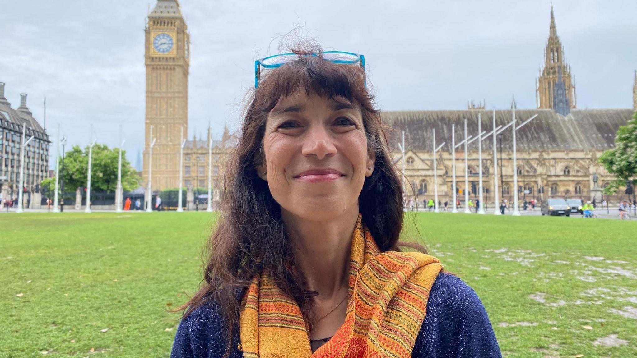 A woman stands in front of the Houses of Parliament. She has brown hair, a blue jumper and an orange scarf. She has glasses perched on her head.