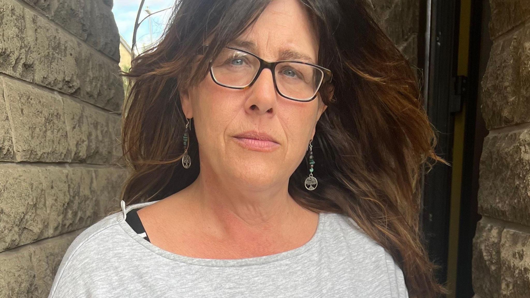 A woman with long brown hair stands between two brick walls. She is wearing dark glasses and has long earrings.