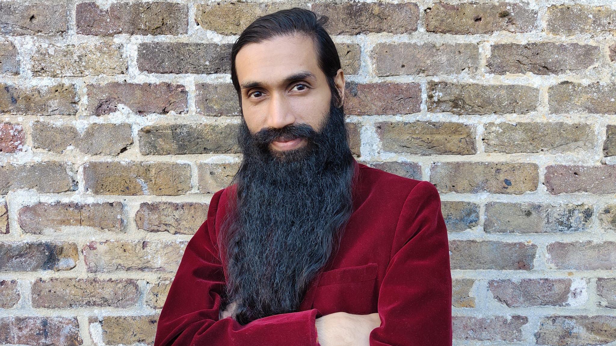 A man with dark black hair and large beard stands in front of a brick wall. He is smiling and wearing a red velvet jacket.
