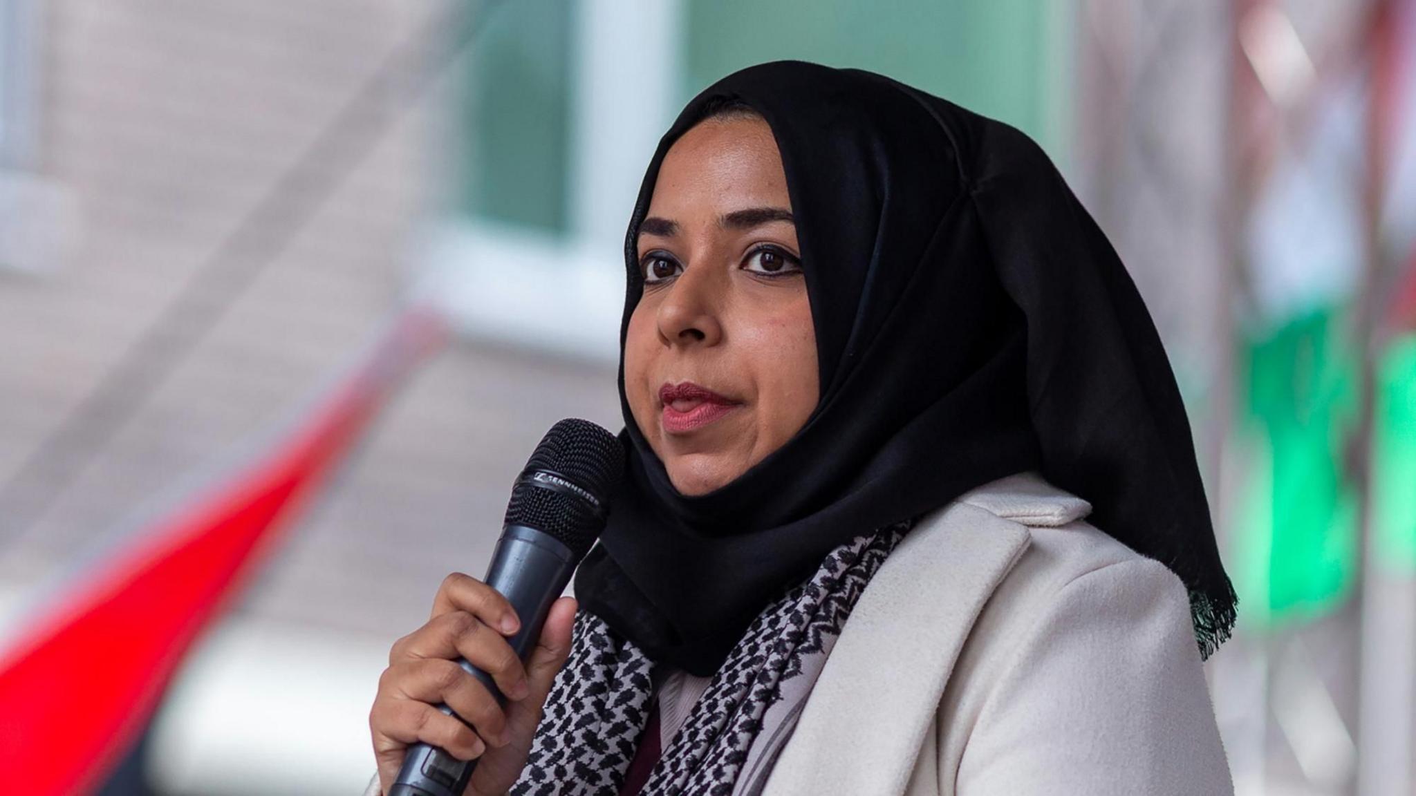 Apsana Begum MP holding a microphone at a demonstration. She is wearing a black hijab and has a patterned black scarf with a cream coat. There is a building and out-of-focus flags behind her.