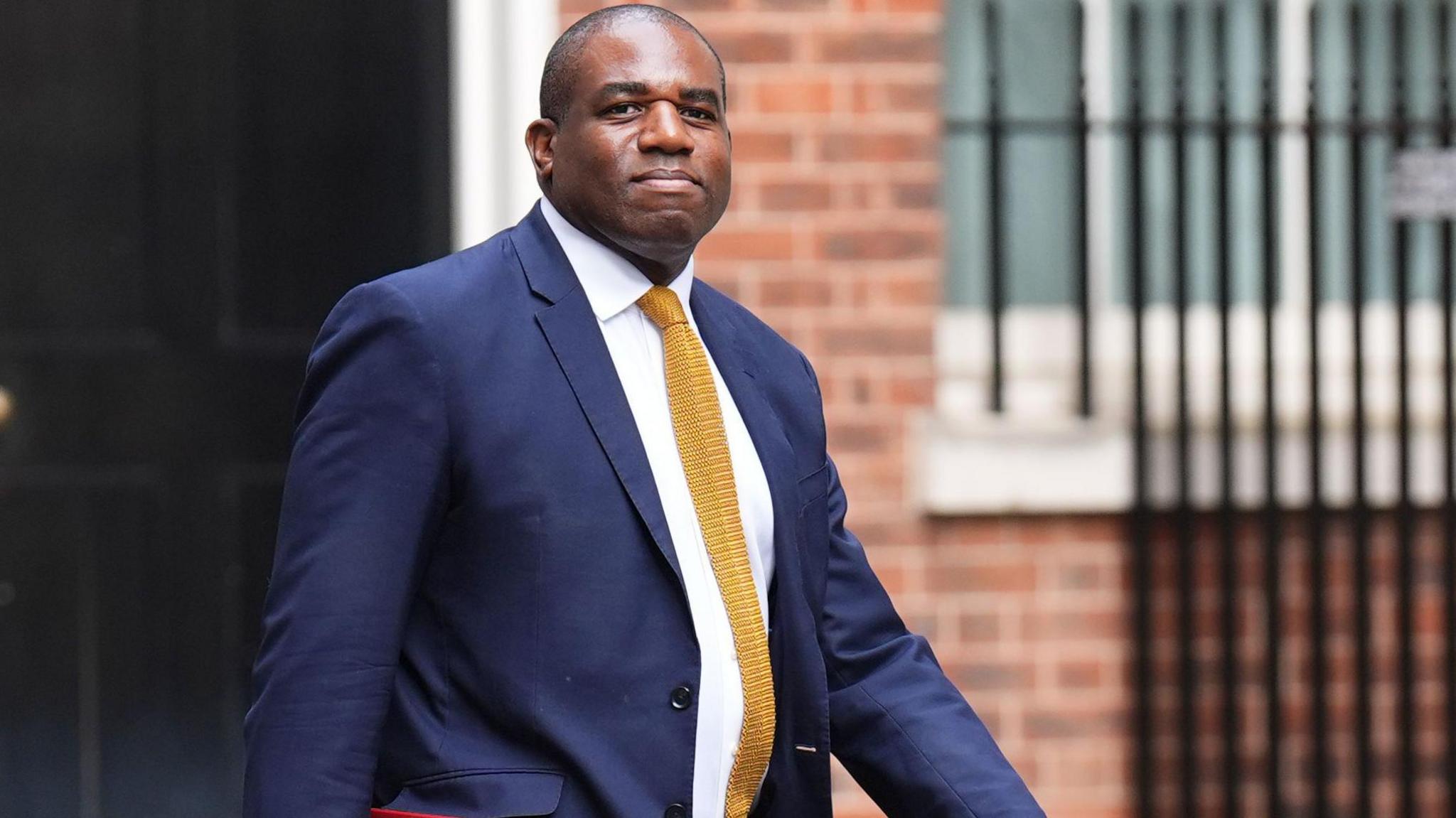 David Lammy walks along in front of a brick building. He is wearing a navy suit and yellow tie while holding a red file.