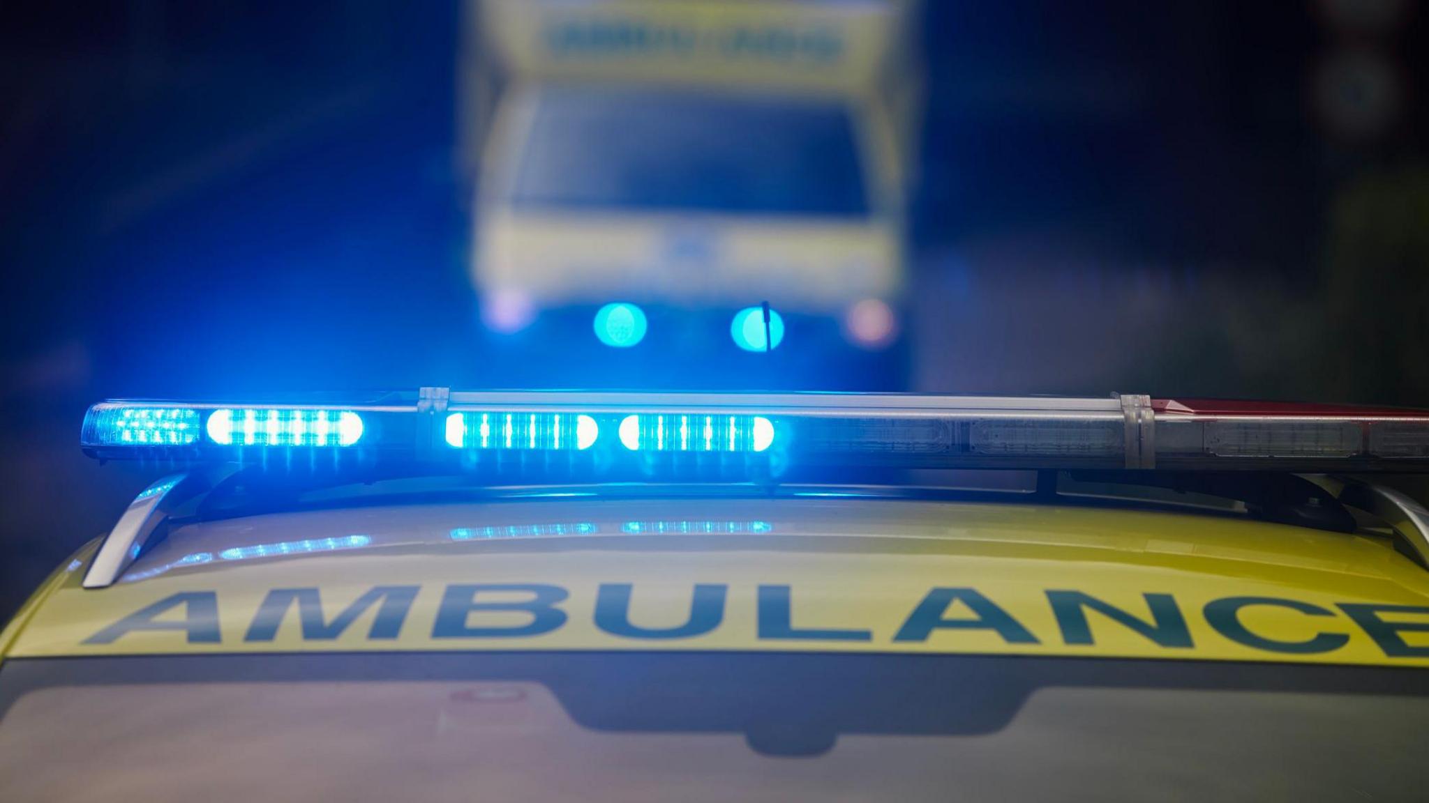 Two green and yellow ambulances in succession with their blue lights on. 