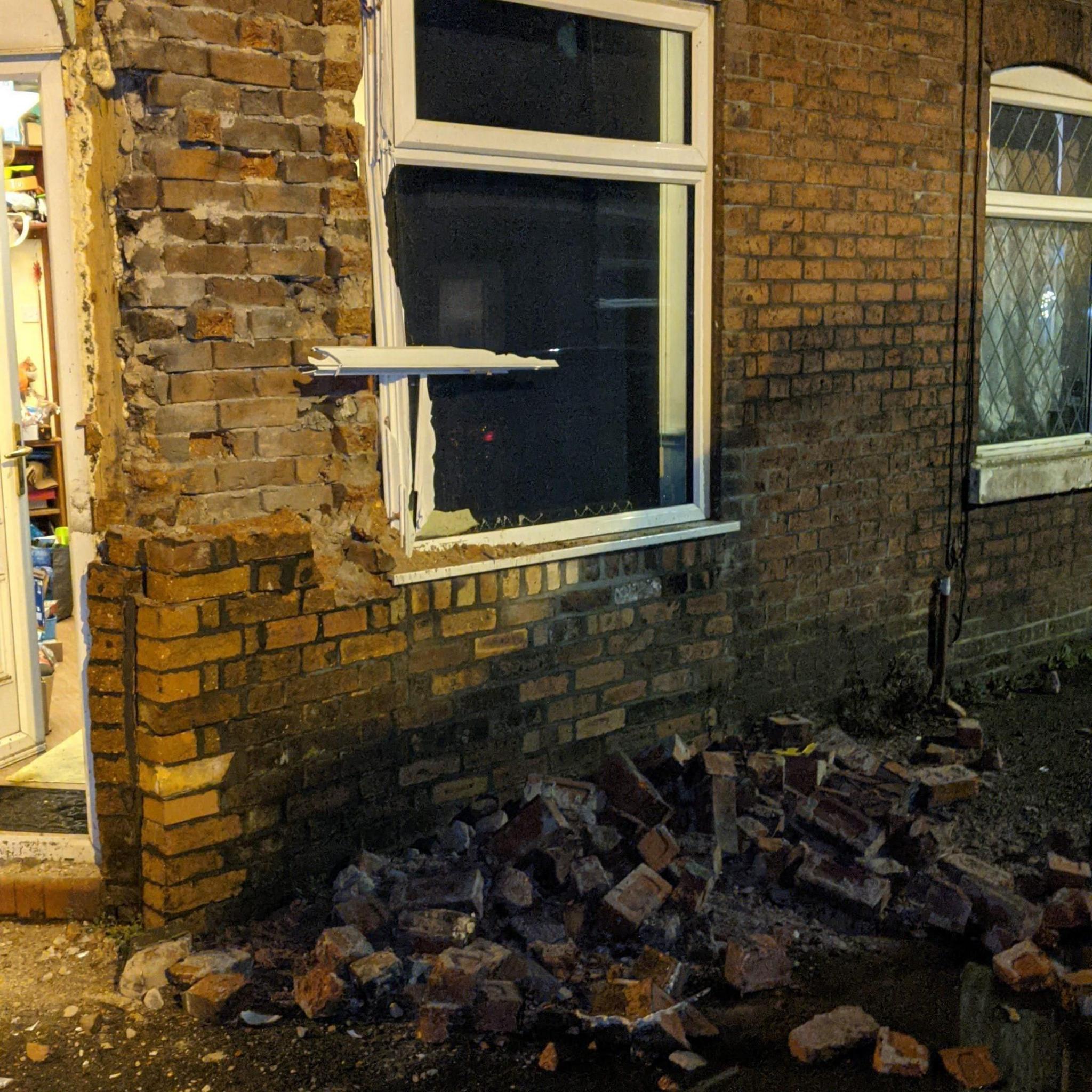 The outside of a house on the corner of a street. The wall appears to have been severely damaged and there are bricks lying on the pavement. The window frame has also been damaged.