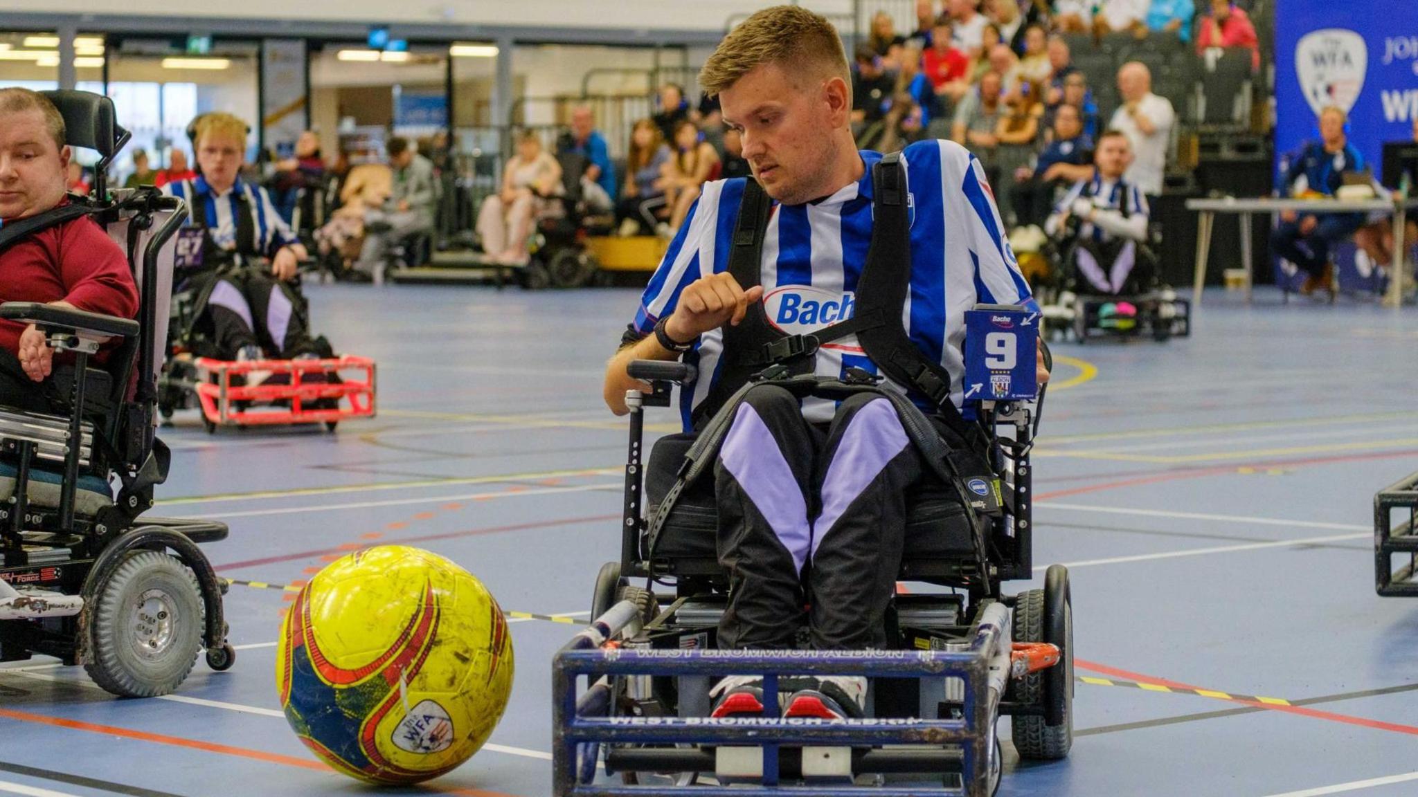 Powerchair Football player coach Chris Gordon in action. He is wearing his Albion kit of white and blue and there is a football in front of his chair. We can see spectators behind him cheering the players on. There are some other players behind Mr Gordon.