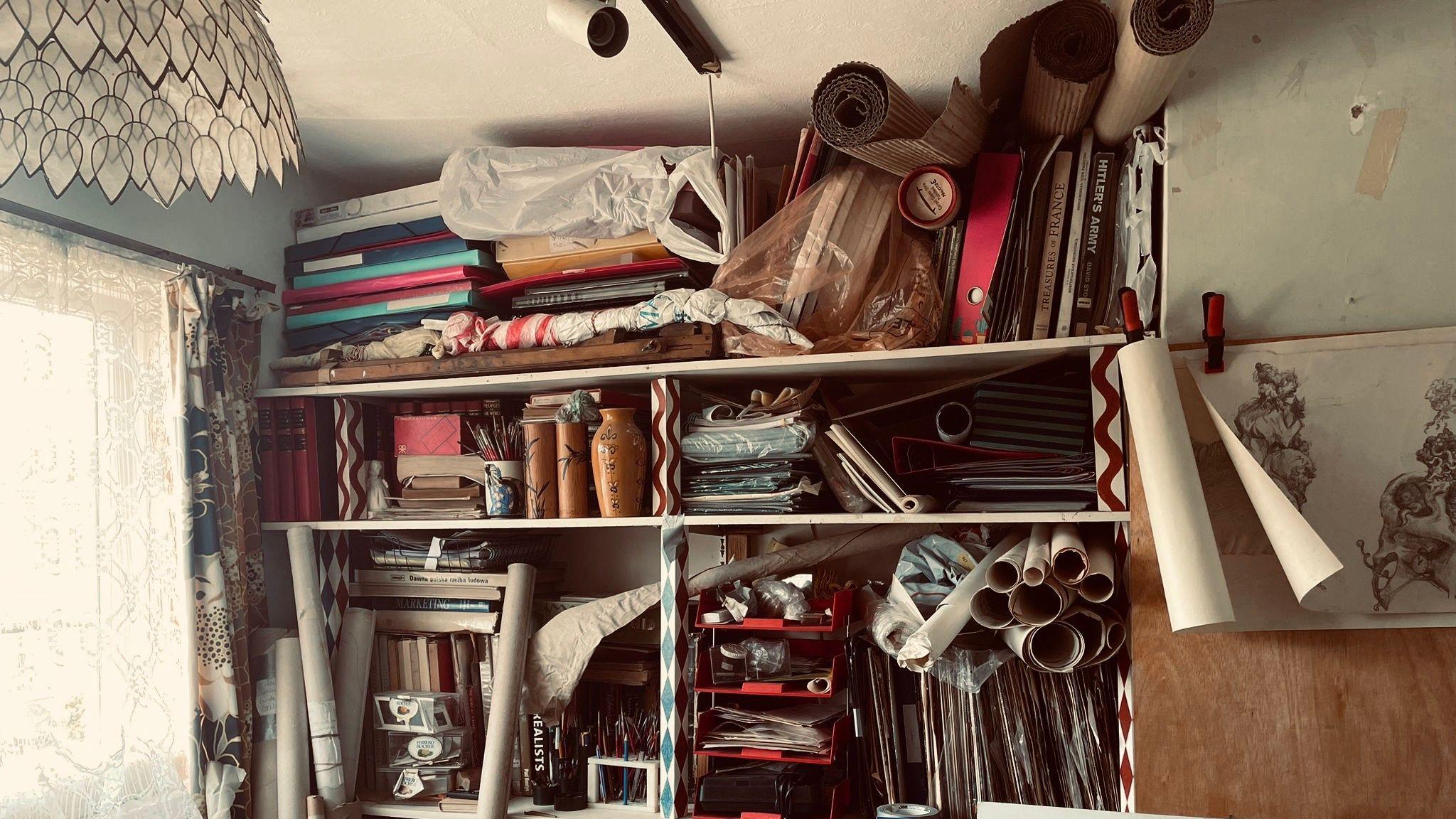 A room with low light showing shelving with rolls of canvases, boards with sketching and folders of paper