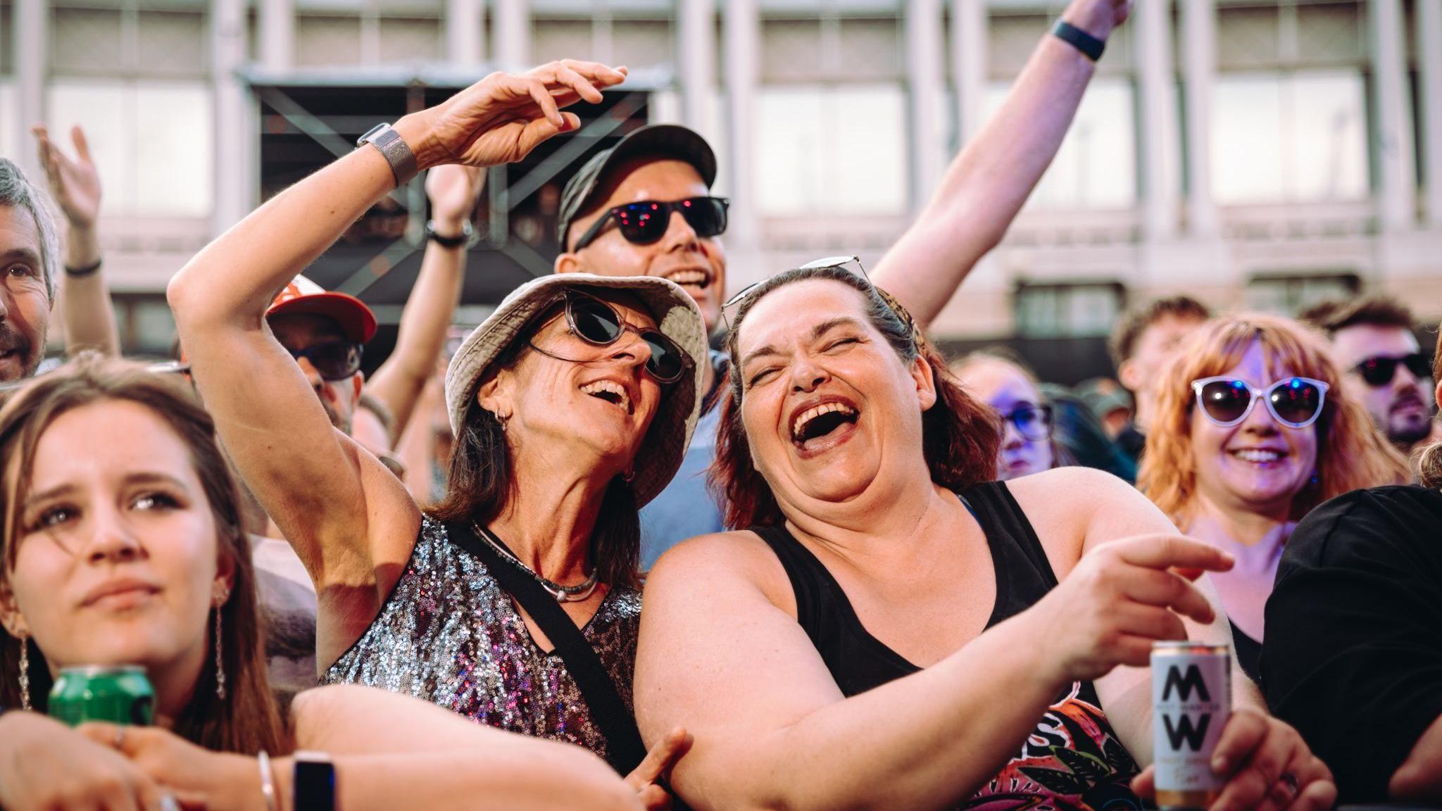 Two women in a crowd at Bristol Sounds. They are both smiling with their mouths open. One of them has her arm up in the air. Other audience members can be seen by them. Many of them are wearing sunglasses, smiling and holding their arms up.