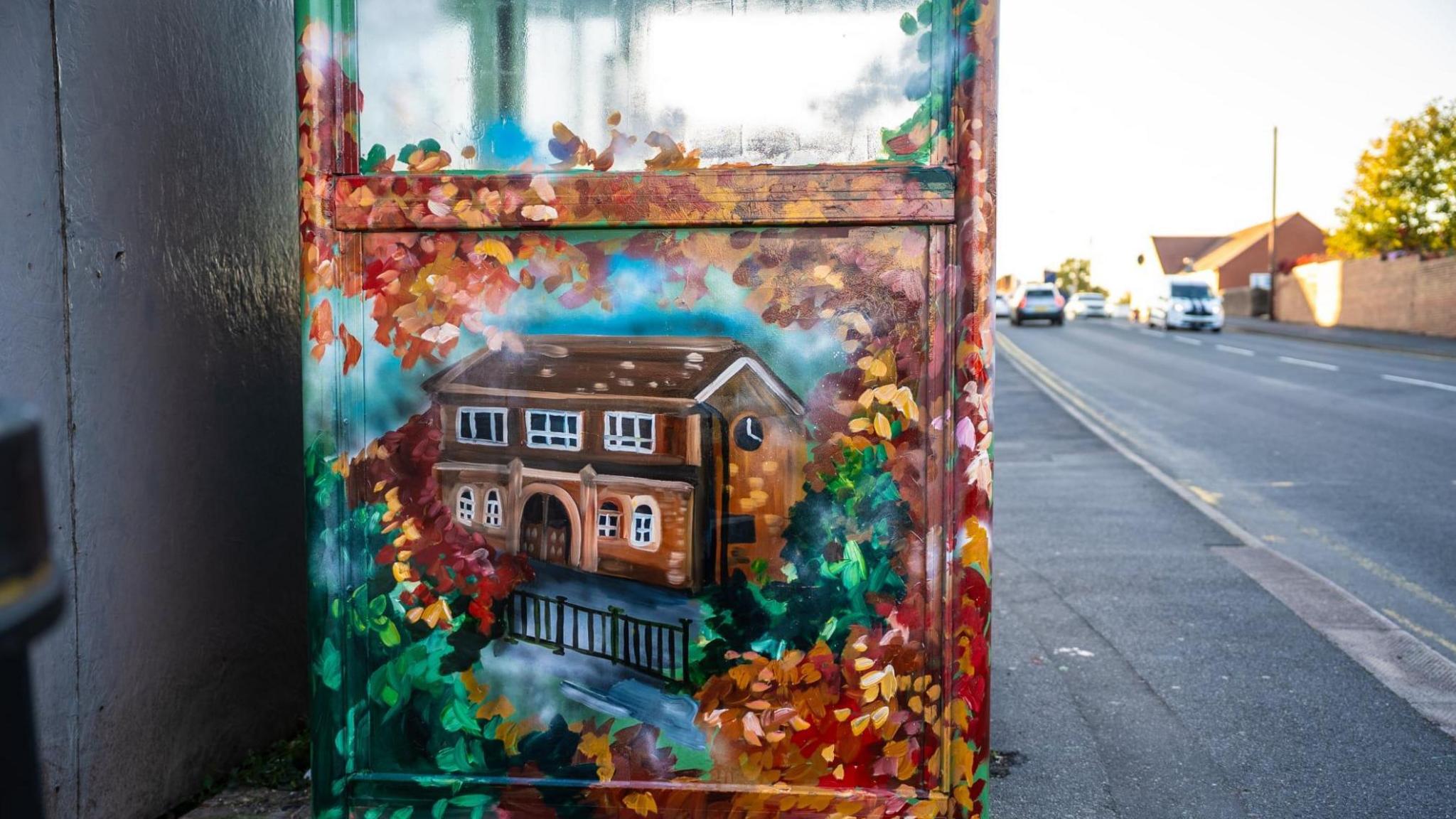 The side of a bus shelter painted with a brown building which has white window frames. Behind it is a blue sky, green foliage and red and brown leaves. 