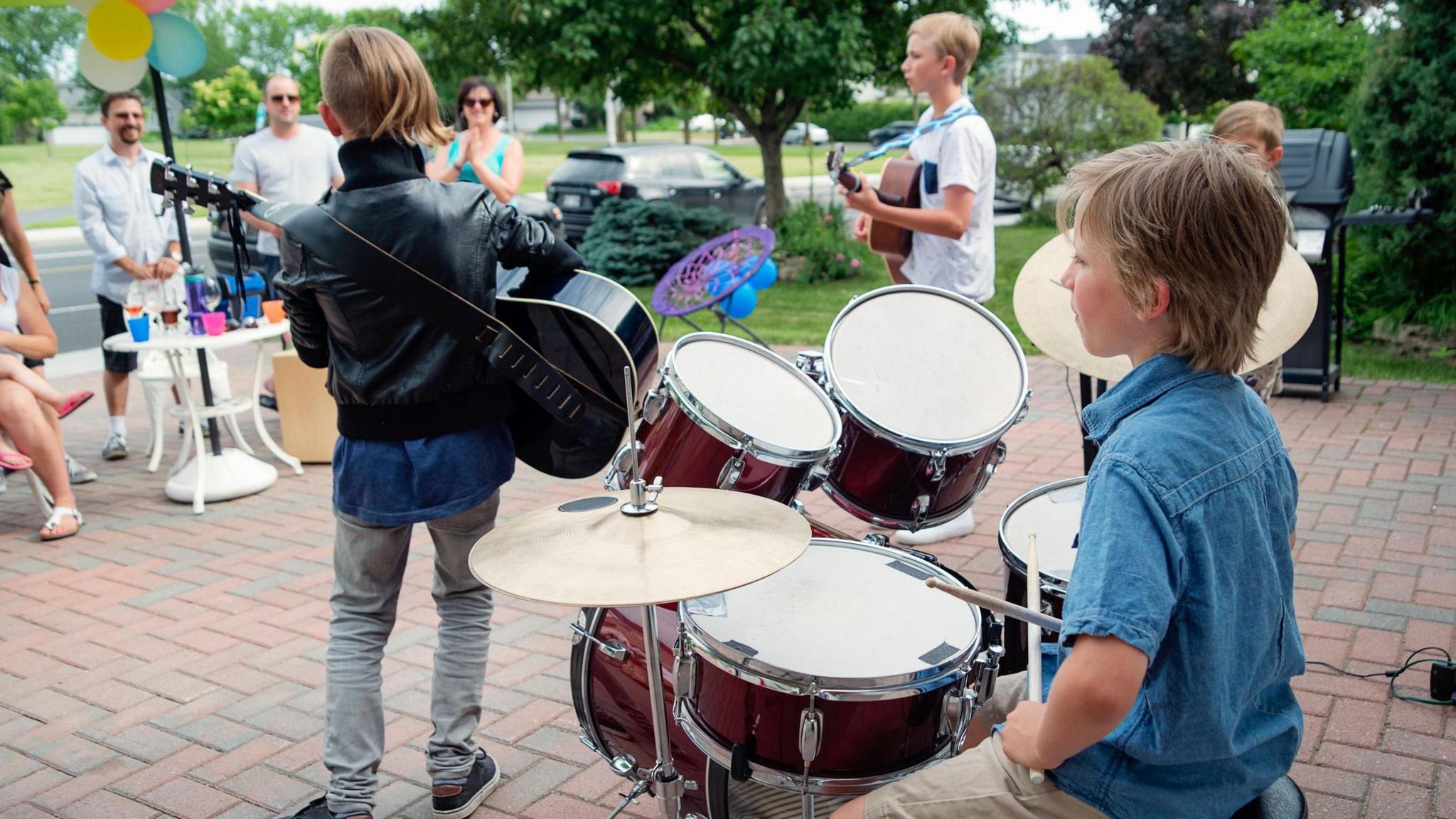 Garage band of young boys between 9 and 12 year’s old, two pairs of brothers, playing in front of family and friends in suburb home driveway on a hot summer day.