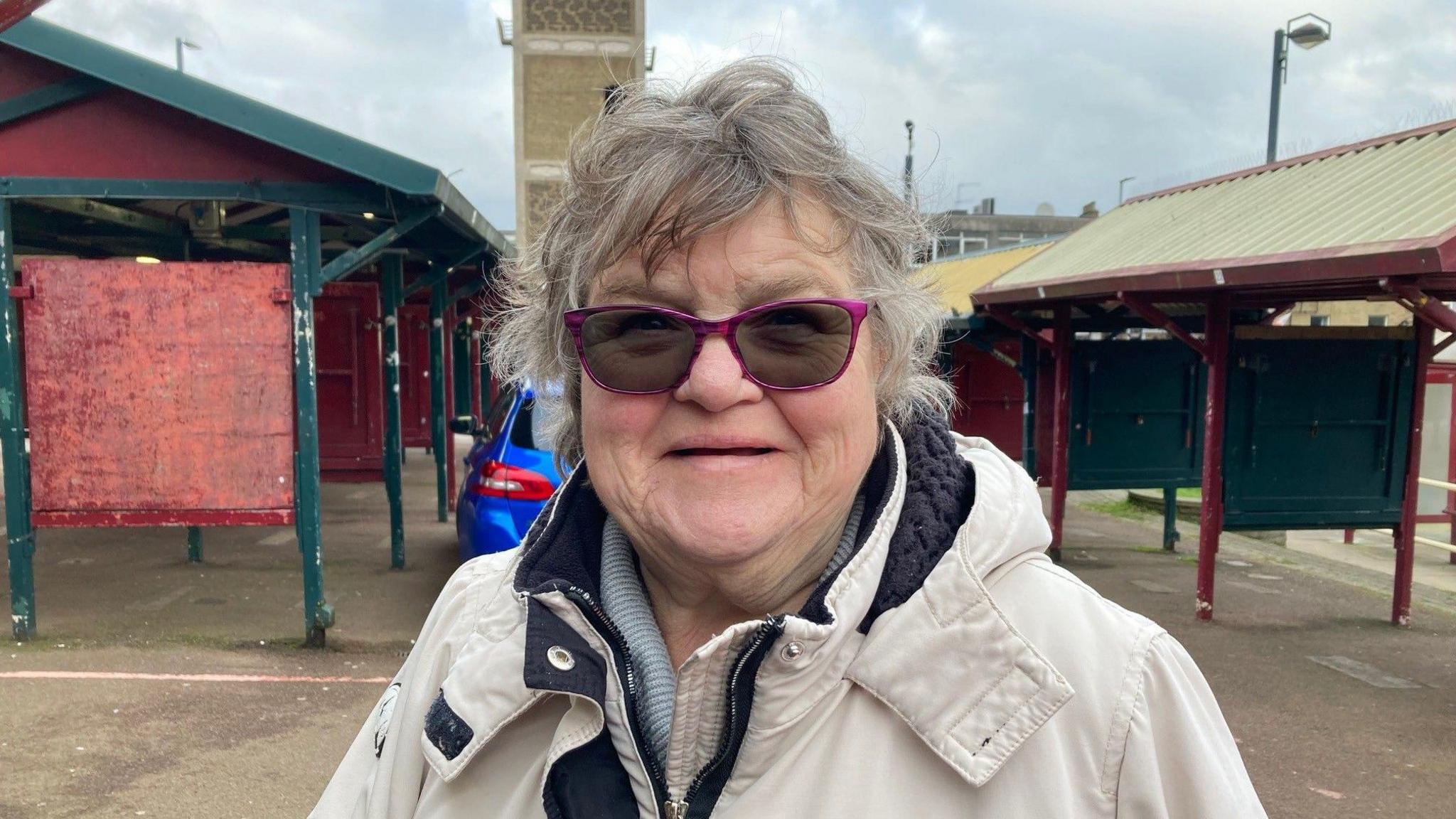 A woman with pink-rimmed glasses and grey hair standing in front of two rows of fixed market stalls.