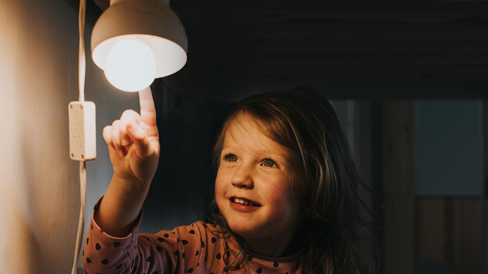 A small girl pointing at a lamp which is switched on. She has blue eyes and dark blonde hair and is smiling at the lamp. 