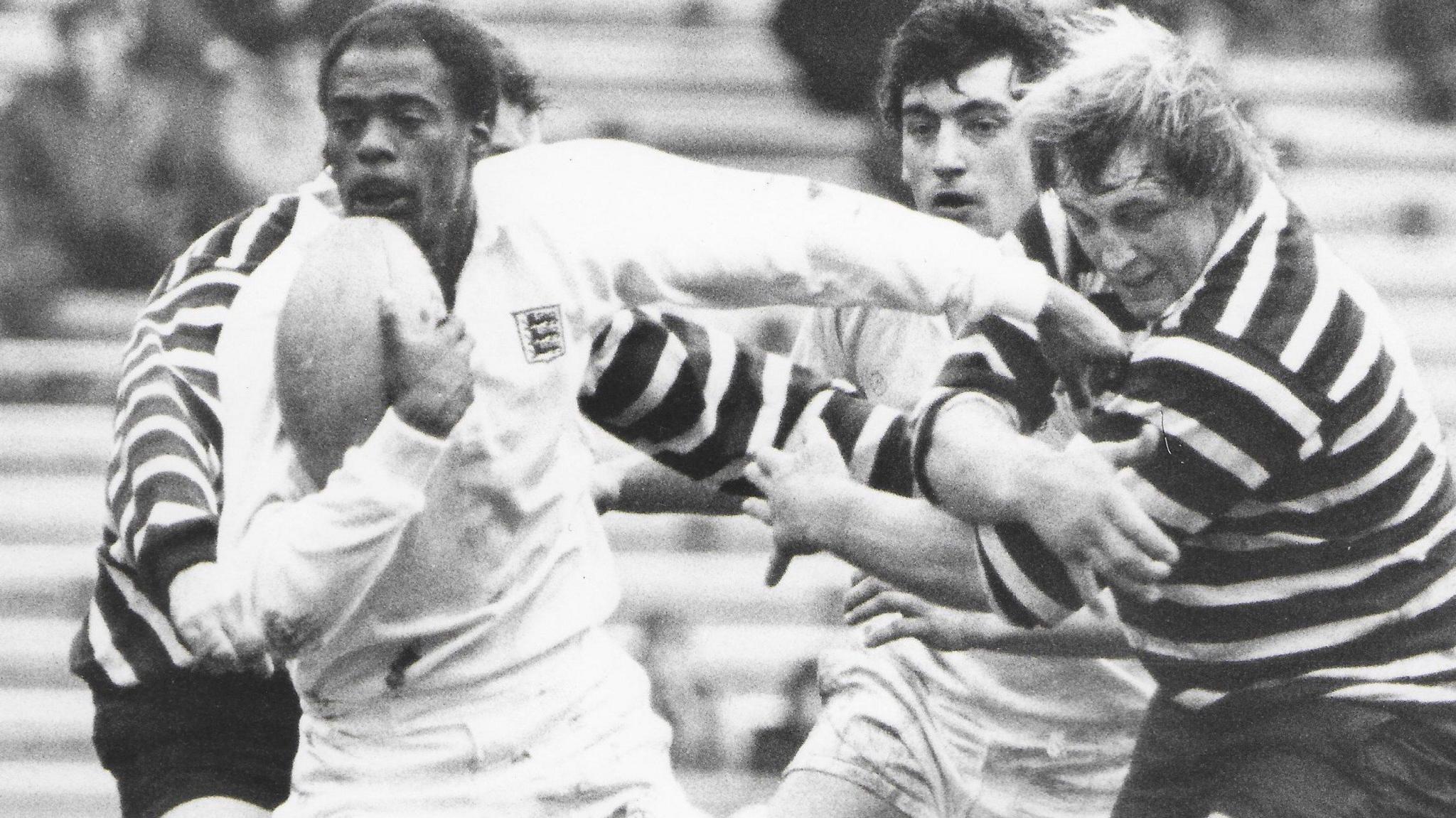 A black and white photo of Ralph Knibbs playing rugby. He is pictured running away with the ball in one hand, with his other arm out in front of another male player.