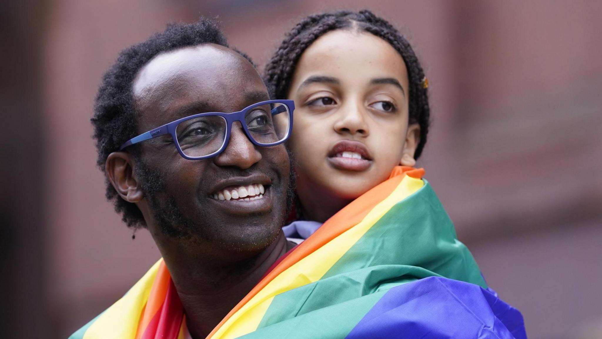 A man wearing a rainbow flag with a child on his back