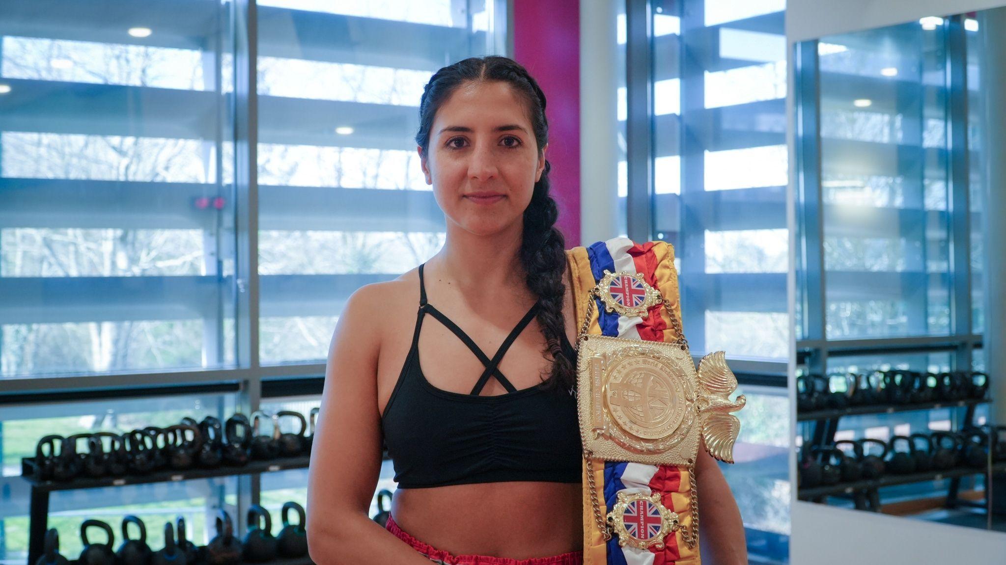 Nicola stands in a gym with a golden sash draped over one shoulder. She has hair hair braided into two french plats and is wearing a black sports bra and red shorts.