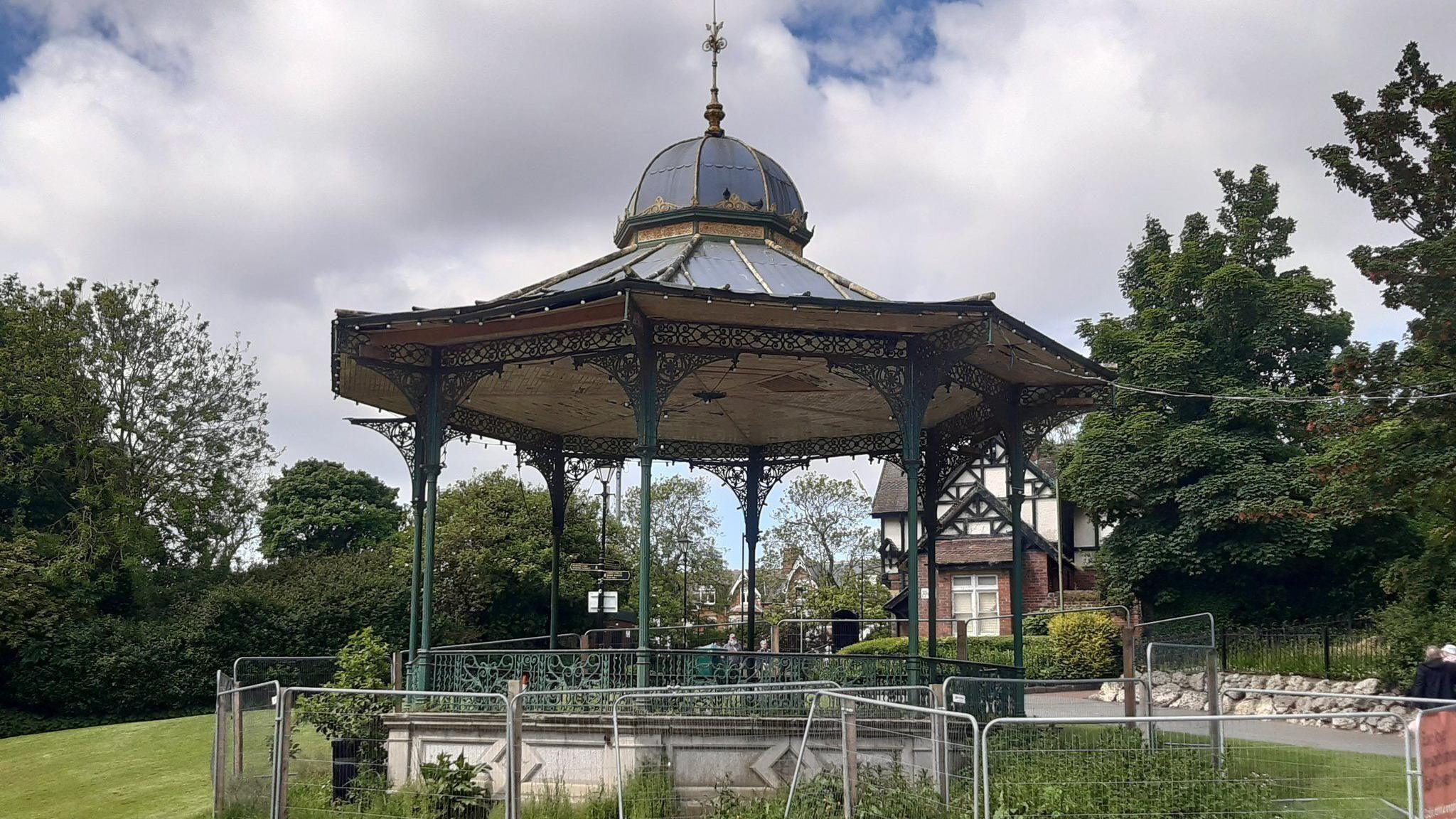 A traditional Victorian octagonal bandstand with domed roof and ornate iron work, showing signs of disrepair. Sitting in a park with a Tudor style lodge in the background, the bandstand is surrounded by temporary metal fence panels.