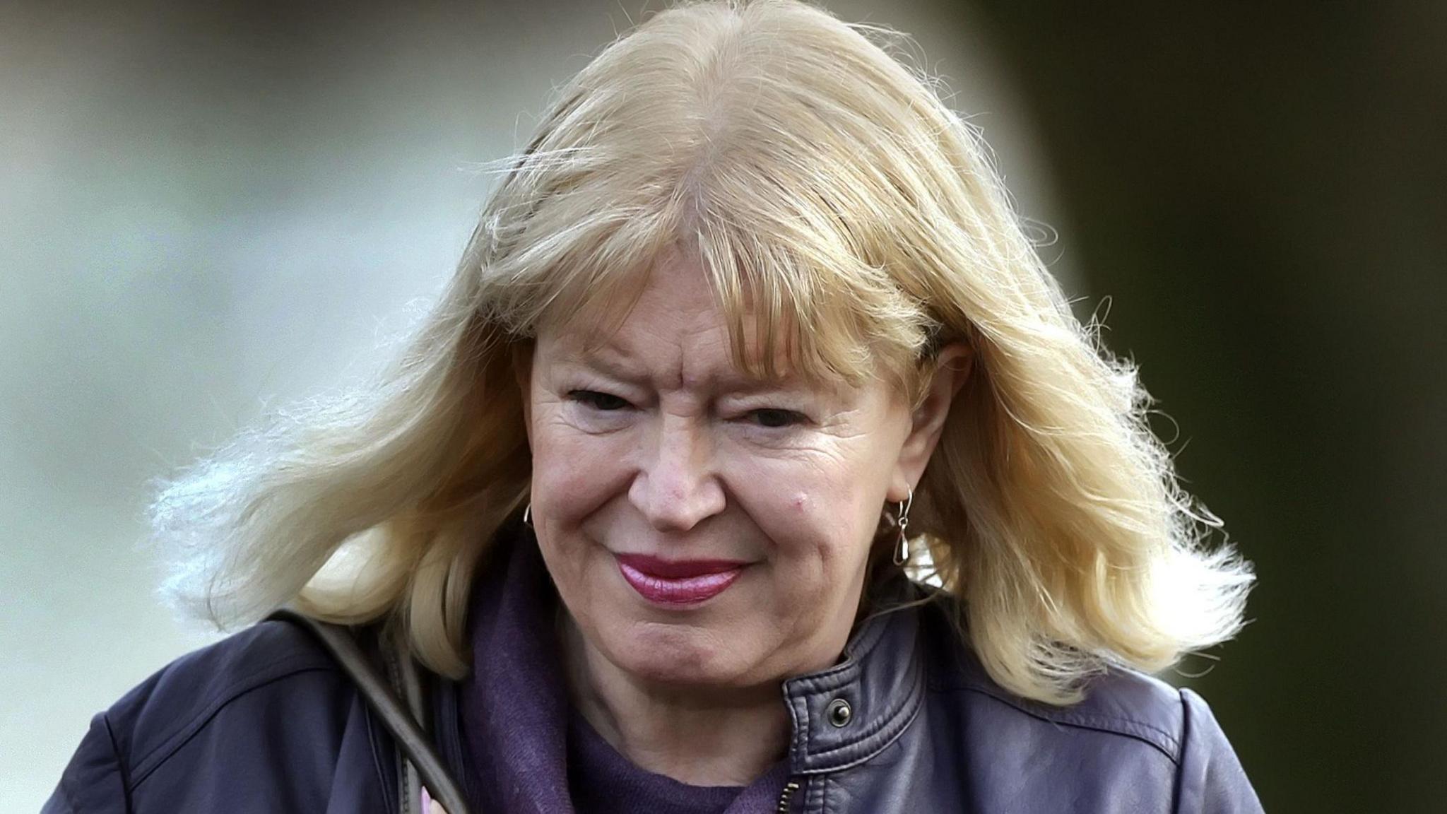 Sandra Brown woman with blonde hair and large earing in a purple leather jacket