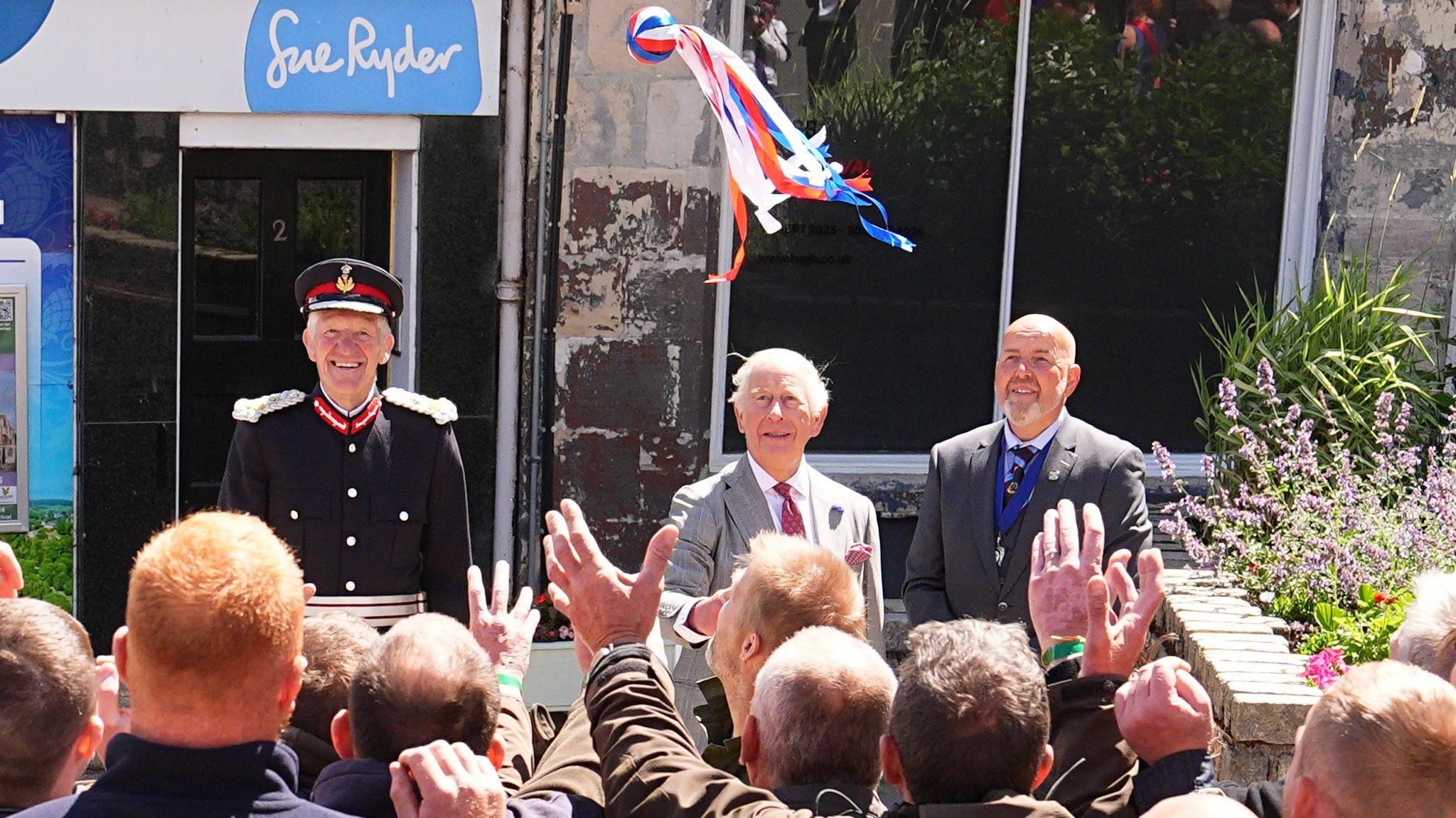 King Charles throws a bright coloured ball with ribbons into a crowd of men waiting to grab it