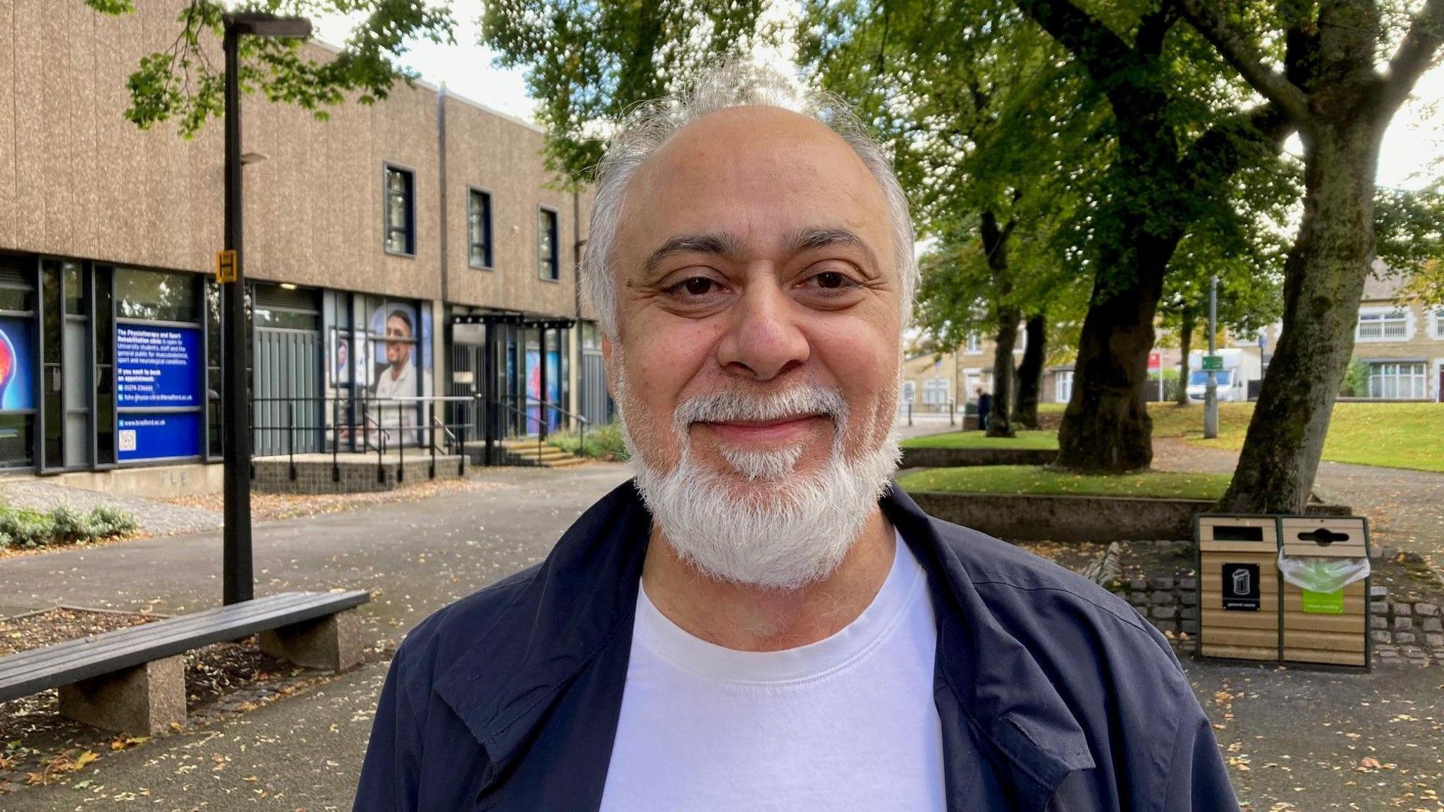 A balding man with white hair wearing a white T-shirt and blue casual jacket. 