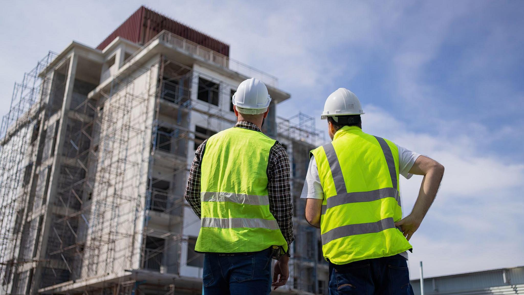 Two men, standing in hi viz vests with white hard hats, facing a large building under construction