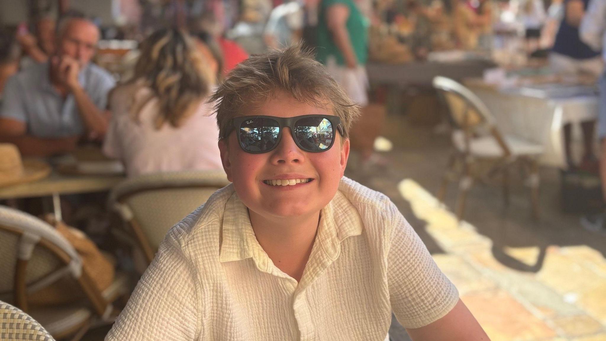 A teenage child wearing a cream shirt and sunglasses, while smiling at the camera.