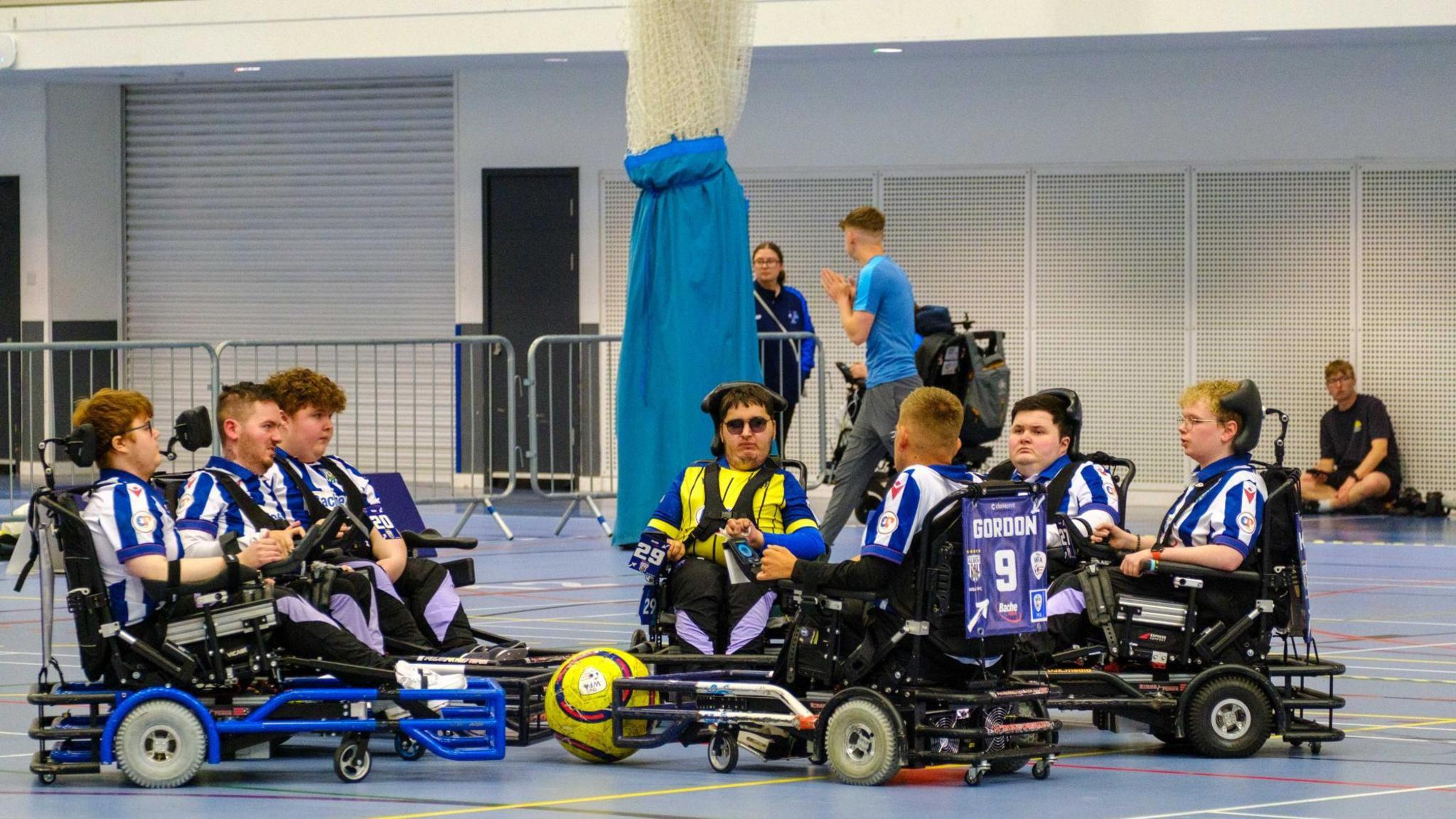 The West Bromwich Albion powerchair football team in a huddle. Six players are on their chairs and surrounding their coach. There is a ball in the middle of them.