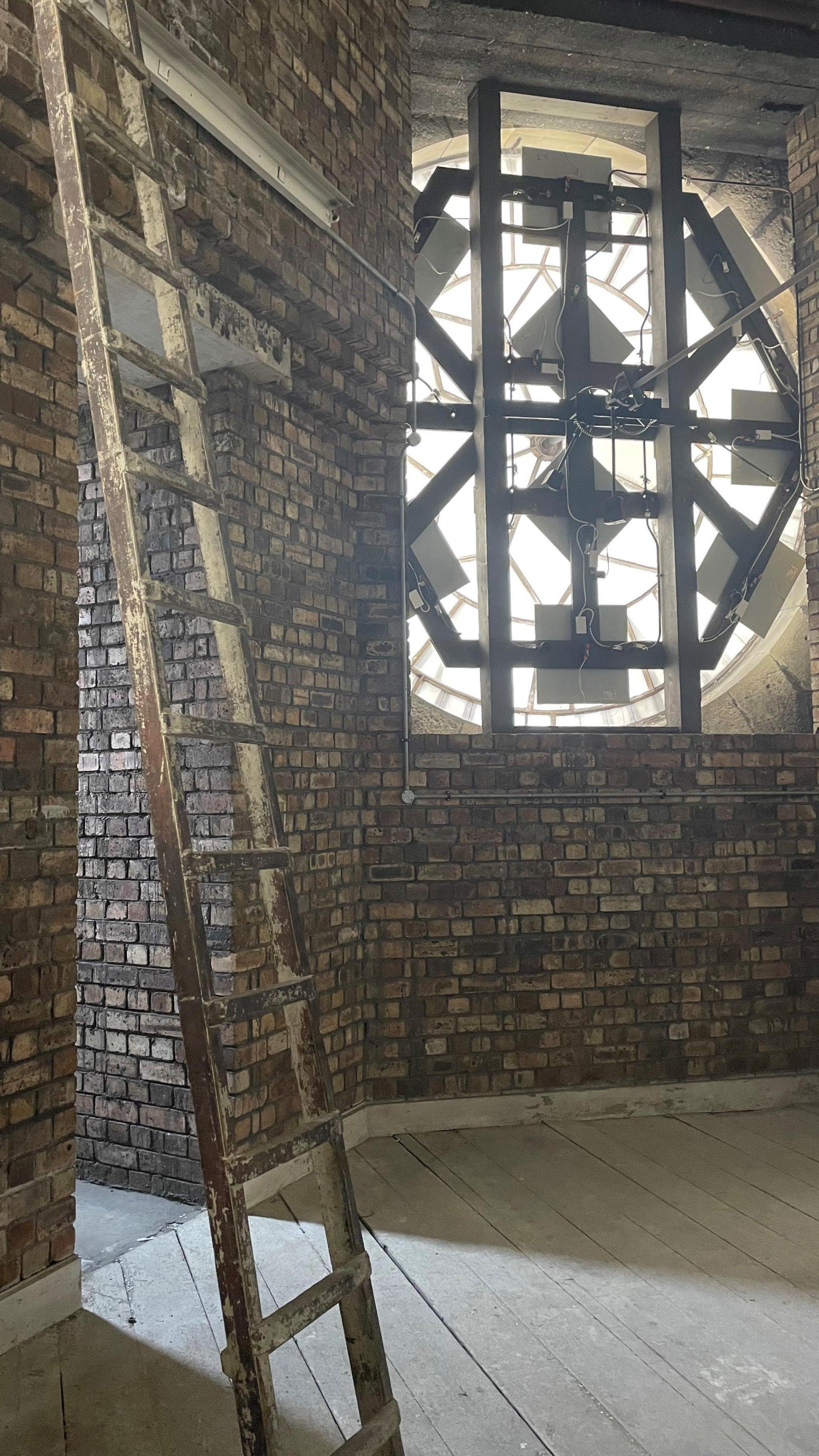 Ten square lights can be seen dotted around the back of the clock face inside the tower. The walls are made of brick and there is a ladder in the foreground.