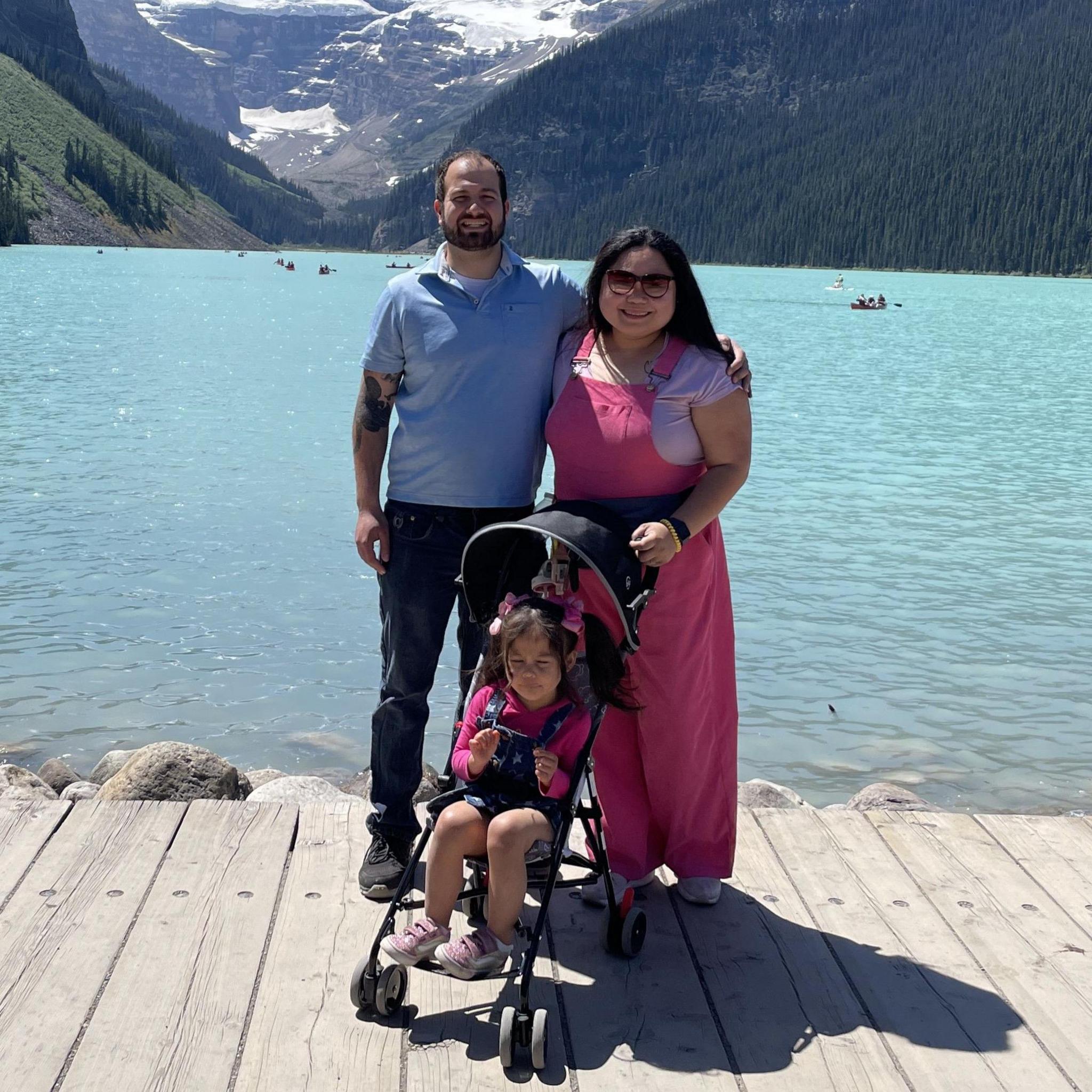 A man and woman stand arm in arm while their daughter sits in a pushchair in front of them. A lake and conifer trees can be seen in the background.