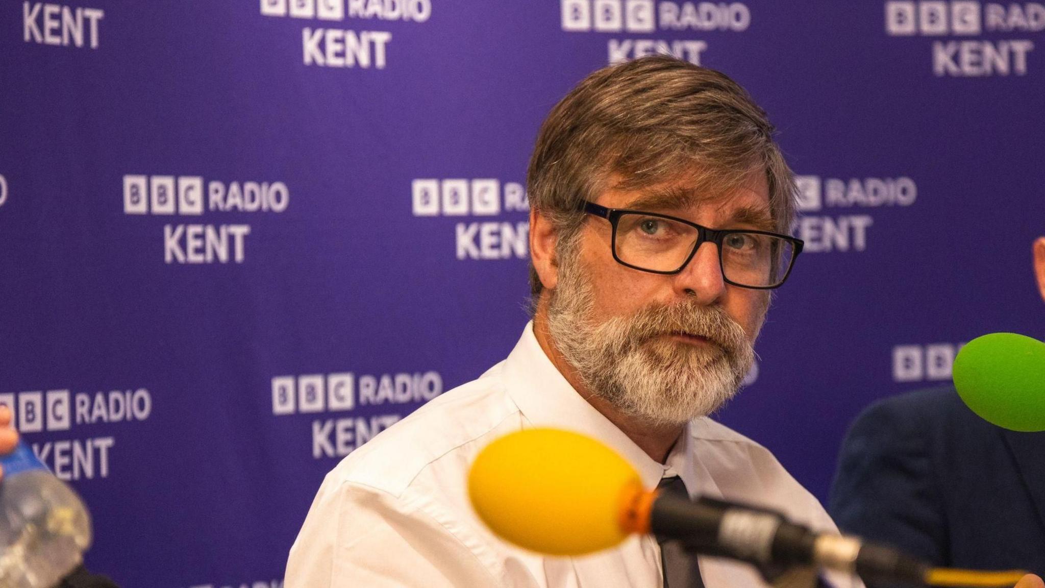 Mark Hood, a man with black glasses and white shirt, answers questions during a BBC Radio Kent political debate. His hair is long, and he has a beard. He is sitting in front of a purple and white backdrop with the BBC Radio Kent logo on it, and has a microphone with a green windsock in front of him.