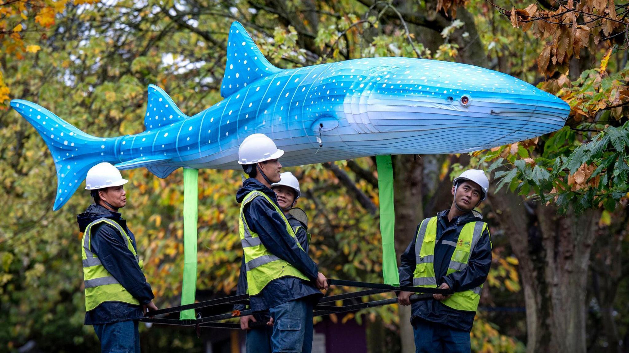 Four men in hard hats and fluorescent jackets carry a lantern shaped like a blue whale above them. Trees can be seen in the background with green and orange leaves