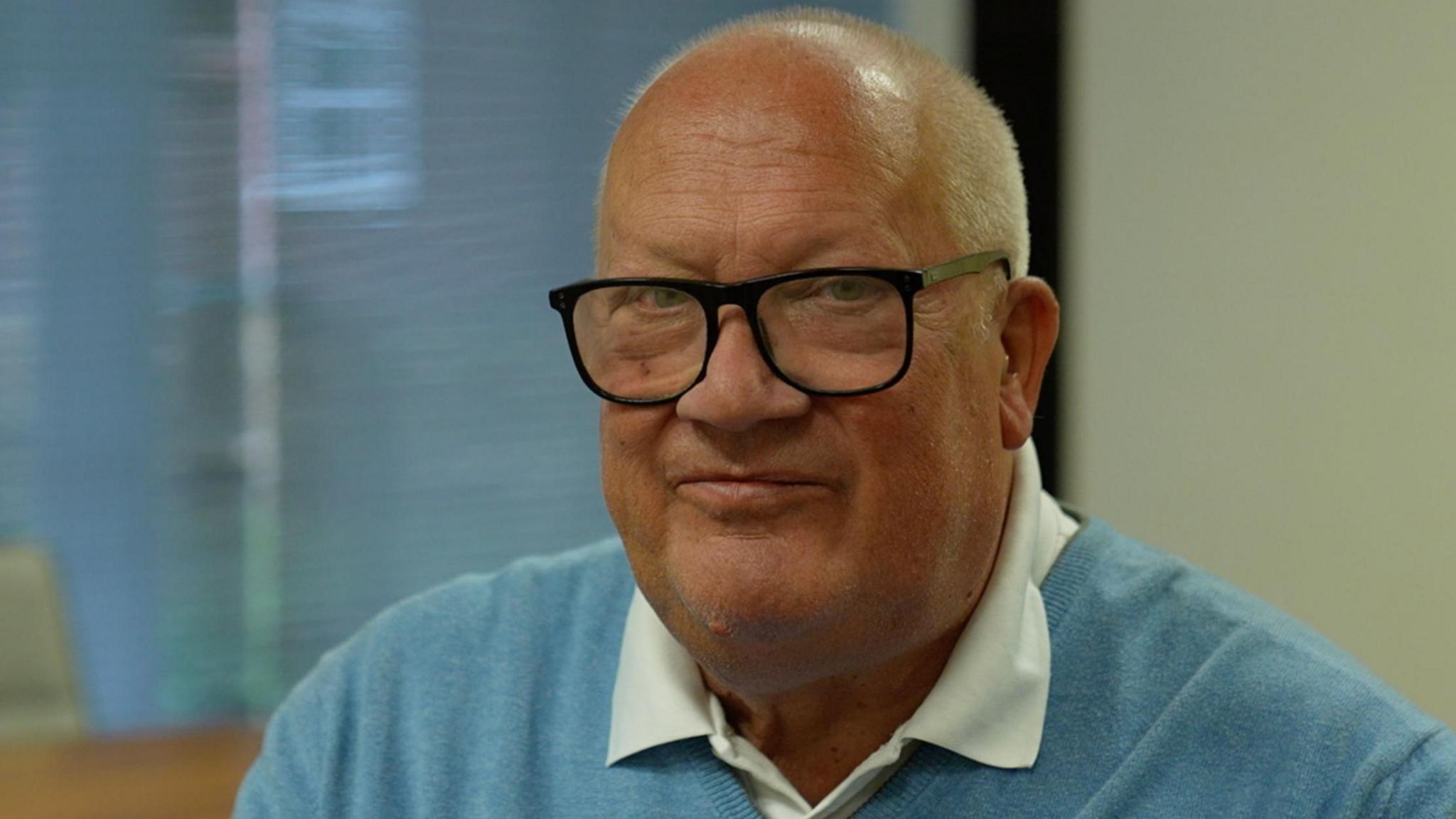 Rob Howard faces the camera wearing black framed glasses, a white shirt and light blue jumper.