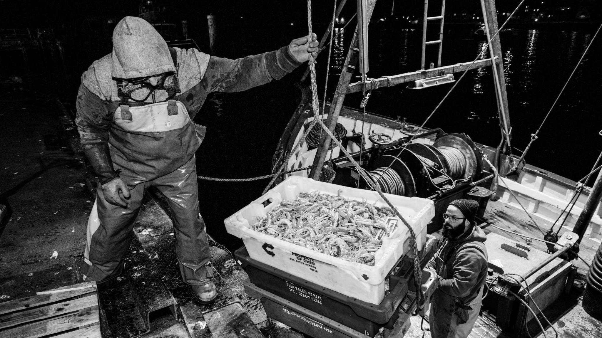 A black and white image of two men landing a catch on the fish quay. One man wearing glasses and a woolly hat is on the boat supporting boxes of prawns as they are hauled up on to the dock. The man on the quayside is wearing waterproof bib and braces.