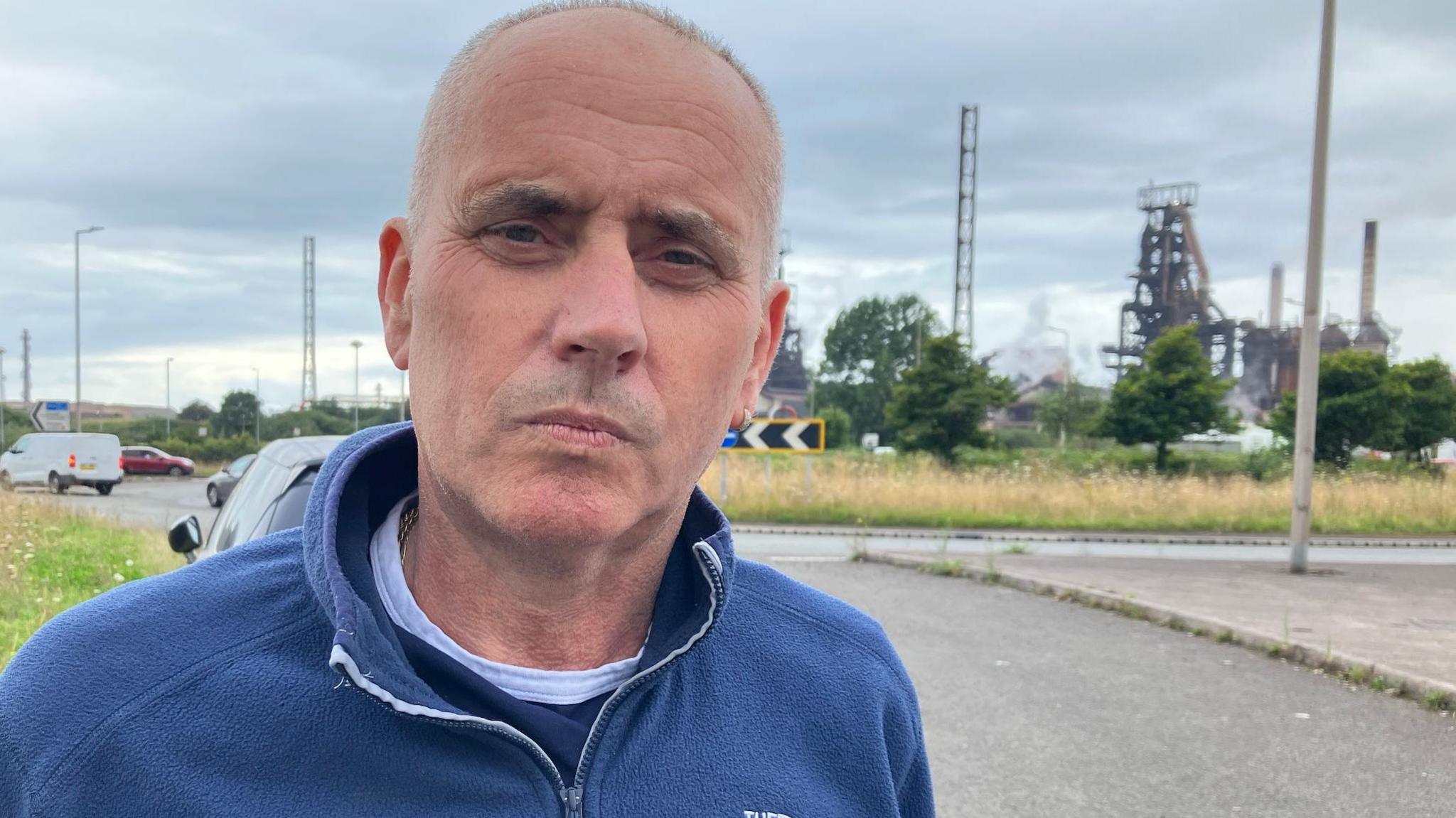 Paul Wagstaffe, wearing a blue jumper standing on the roadside in front of Tata Steel's blast furnaces in Port Talbot, in the background