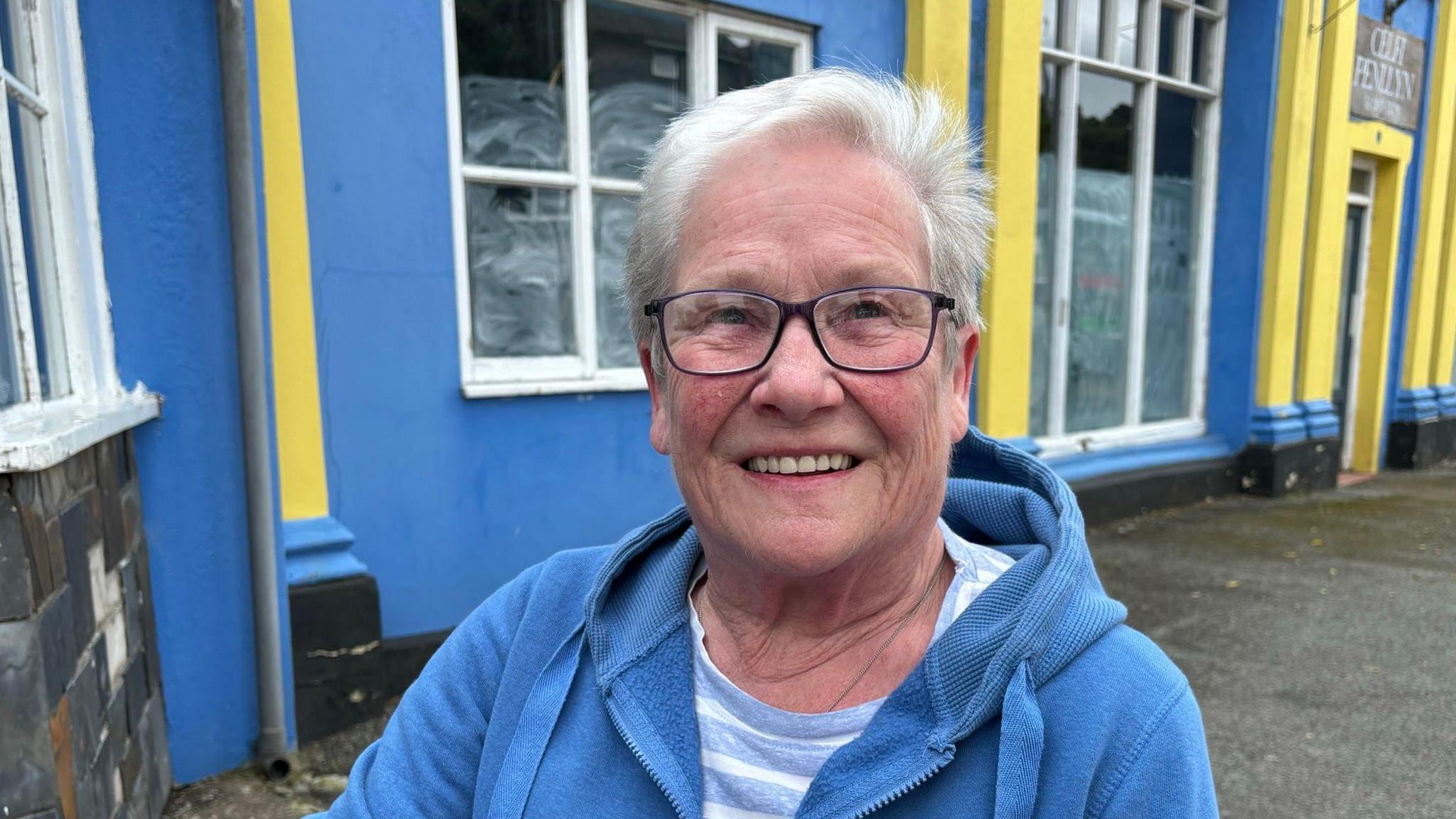 Beryl Roberts stands in front of a blue and yellow house smiling at the camera