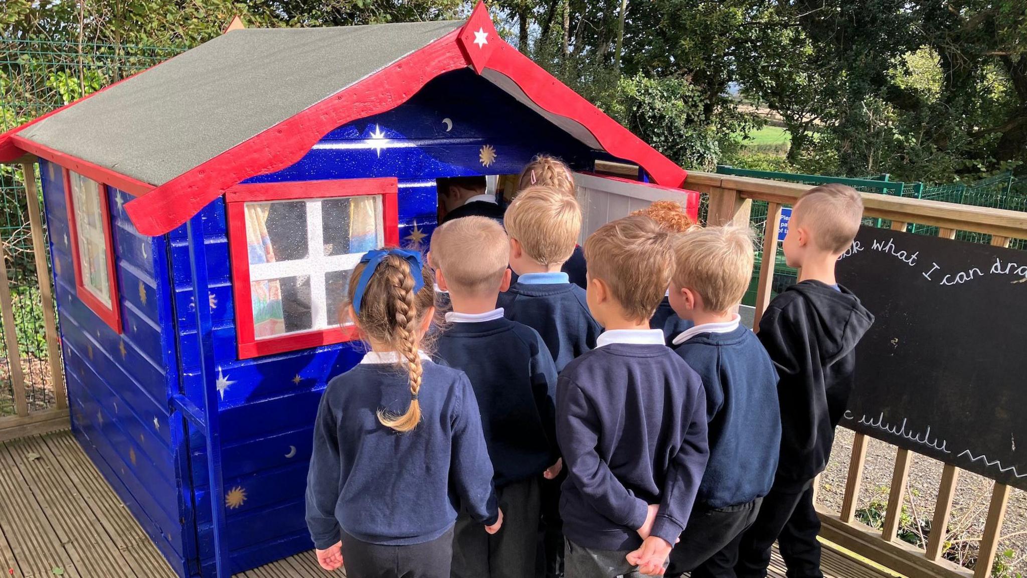 Several primary school children in blue uniforms in front on a blue and red Wendy house waiting to get in.