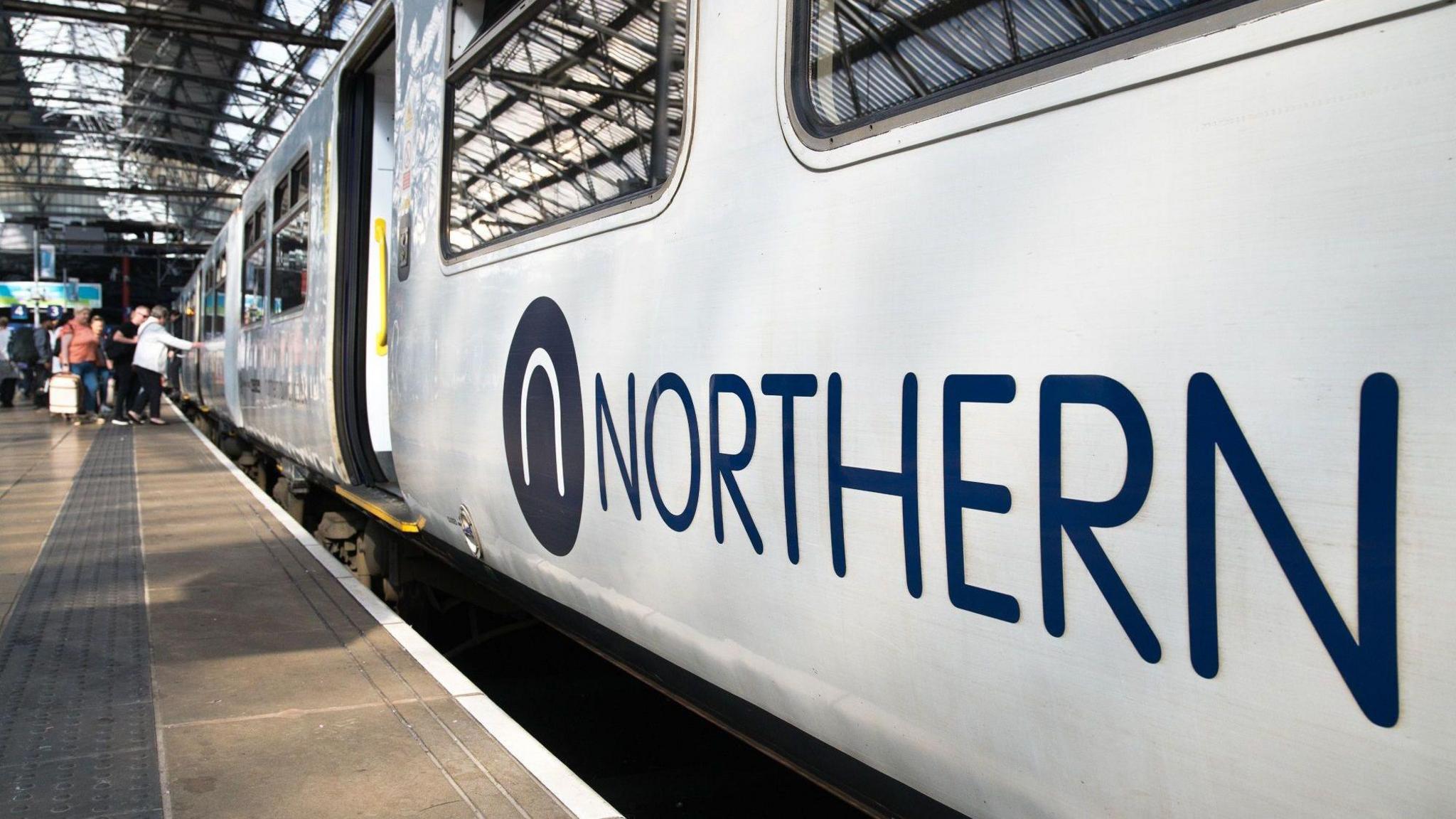 A Northern train is stationery at a platform, with about four people seen preparing to board it. One of the passengers has a wheeled suitcase. A blue sign saying this is platform three can also be seen in the distance. 