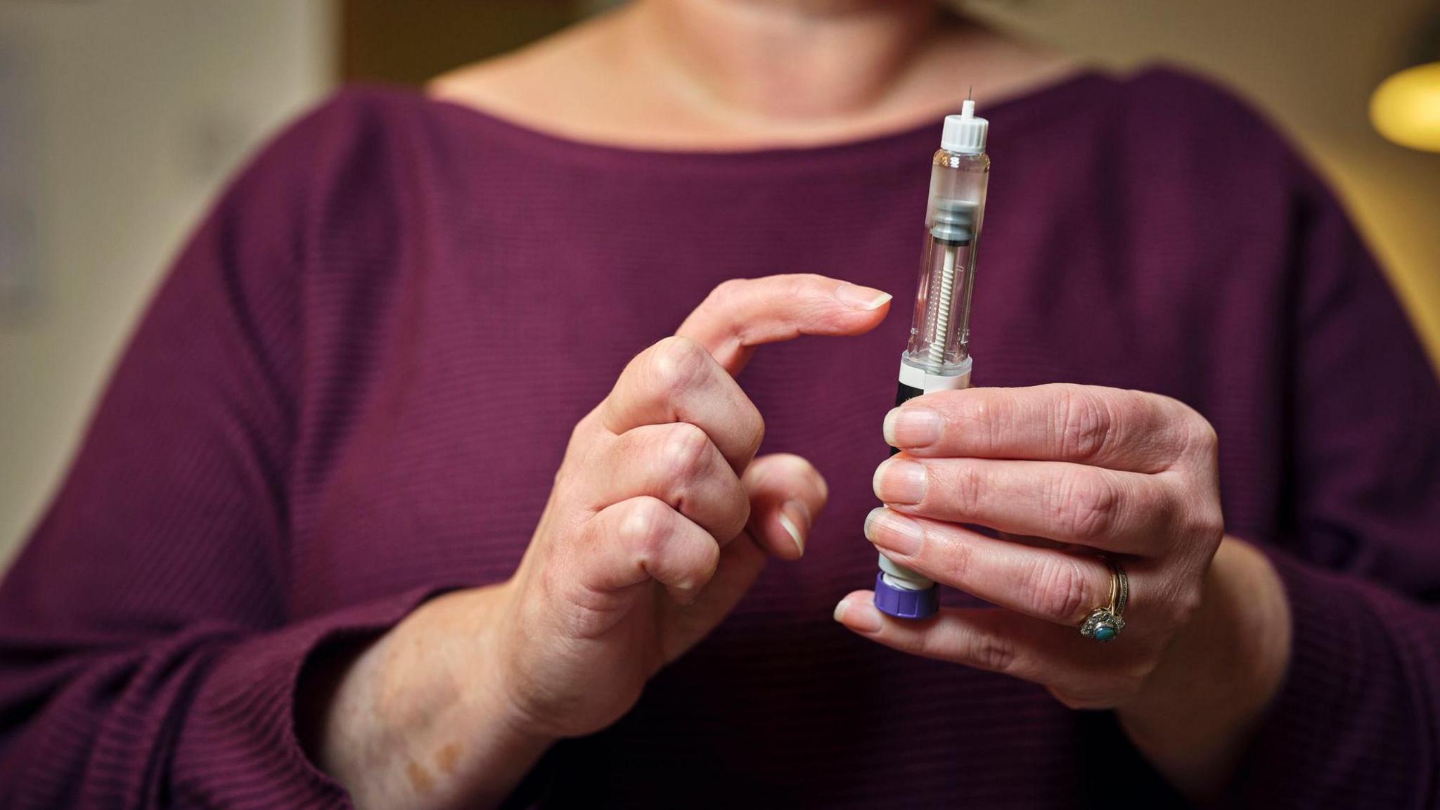 A woman wearing a maroon jumper prepares for a subcutaneous self-injection with a semaglutide pen. She taps it with her right hand and holds it in her left hand.