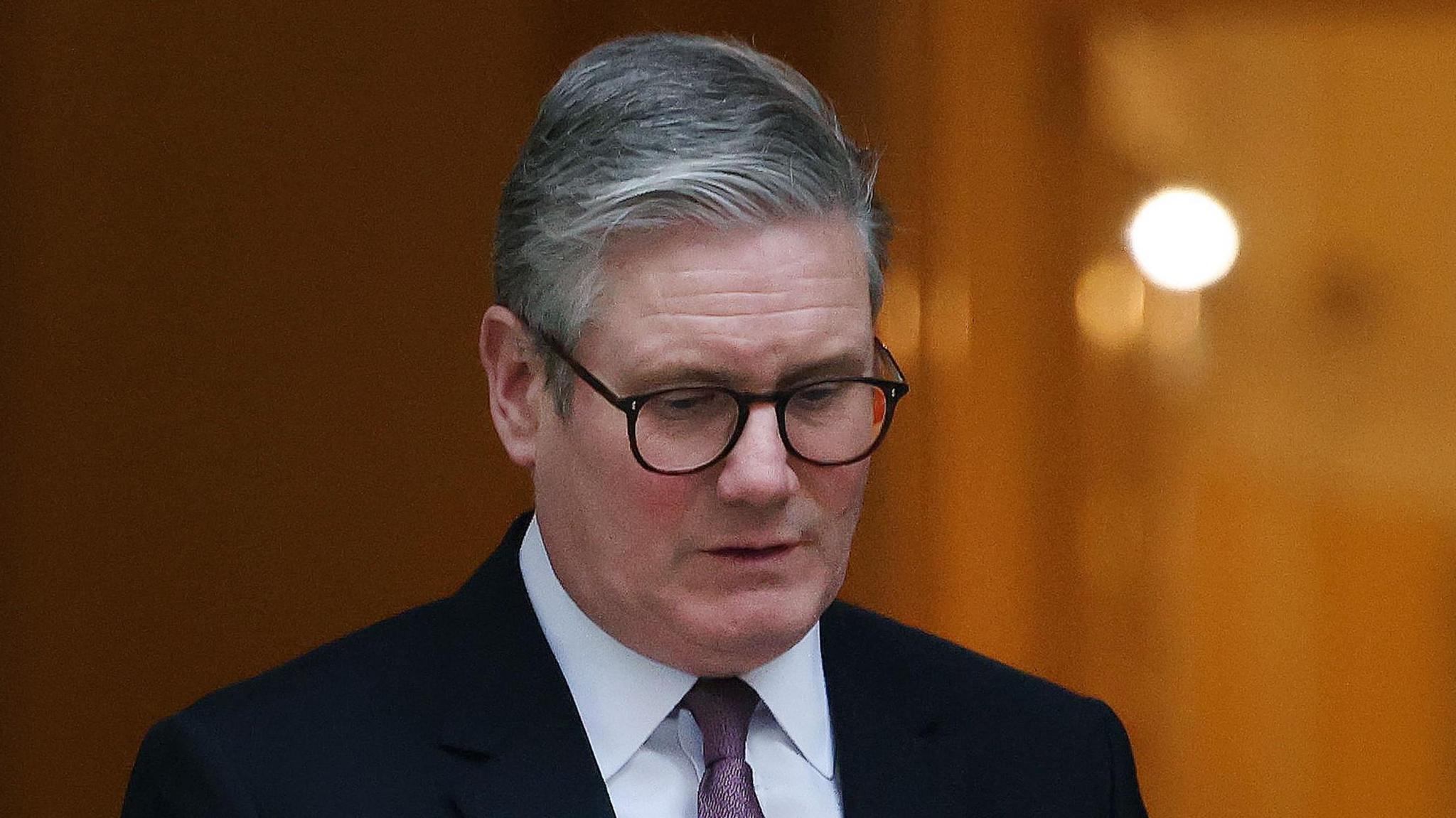 Sir Keir Starmer wearing dark suit and glasses looks down at floor