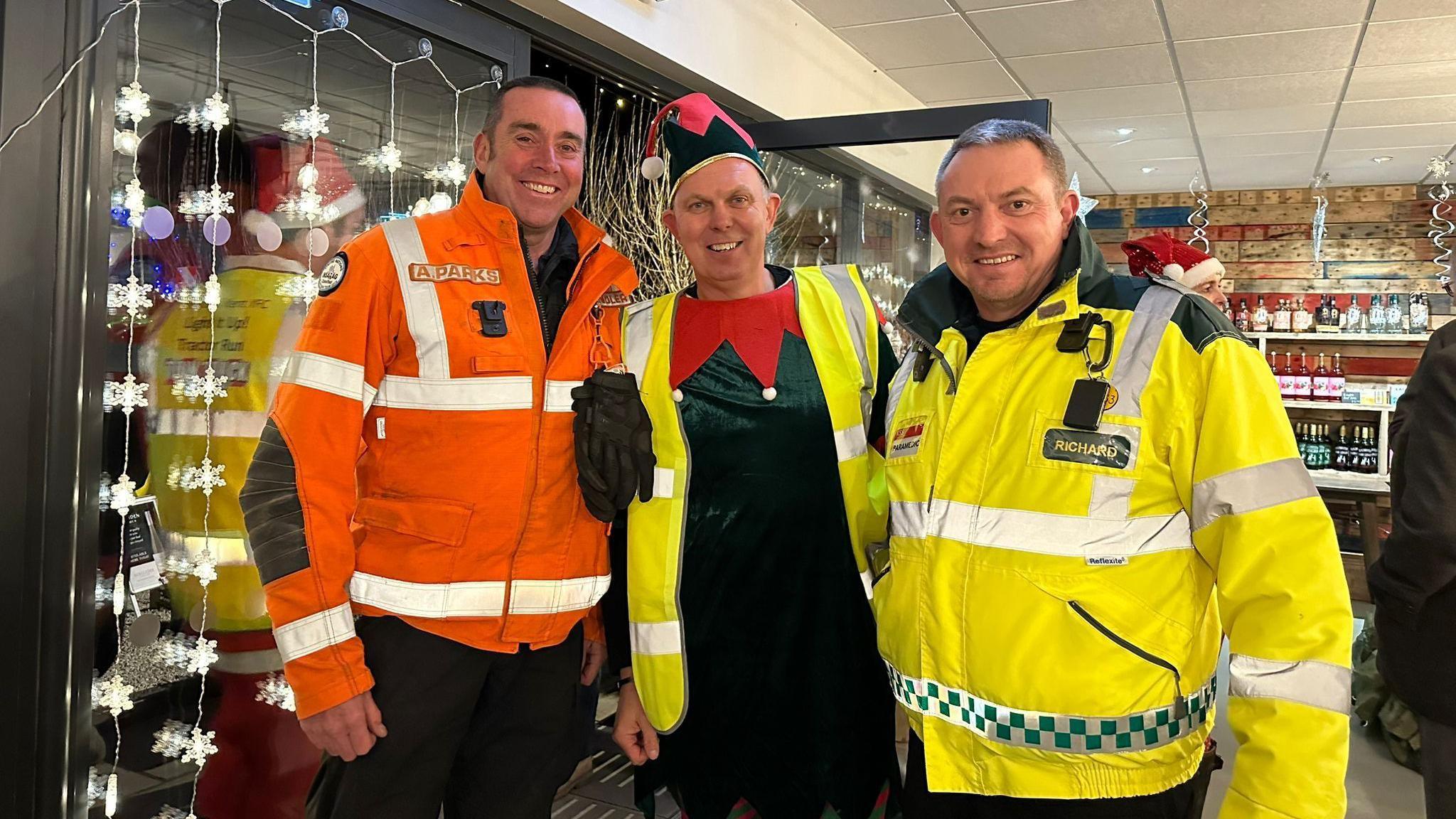 Andy Parks of Kent Fire and Rescue Service, Mark Brown Weald of Kent YFC Club Leader and Richard de Coverly of the Air Ambulance Kent Surrey & Sussex stand together and look at the camera smiling. Andy and Richard are in high vis clothing while Mark is wearing an elf costume with a high vis gilet over the top. There are fairy lights strung up in the background.