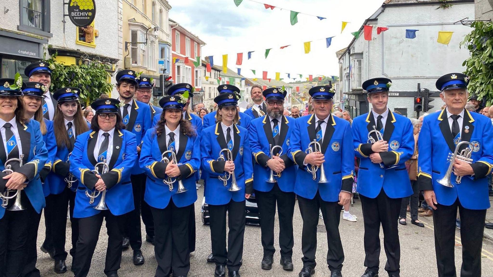 A band wearing blue blazers with black lapels and match ties, as well as matching black caps are seen in the street. Each member of the band pictured is carrying a silver trumpet.