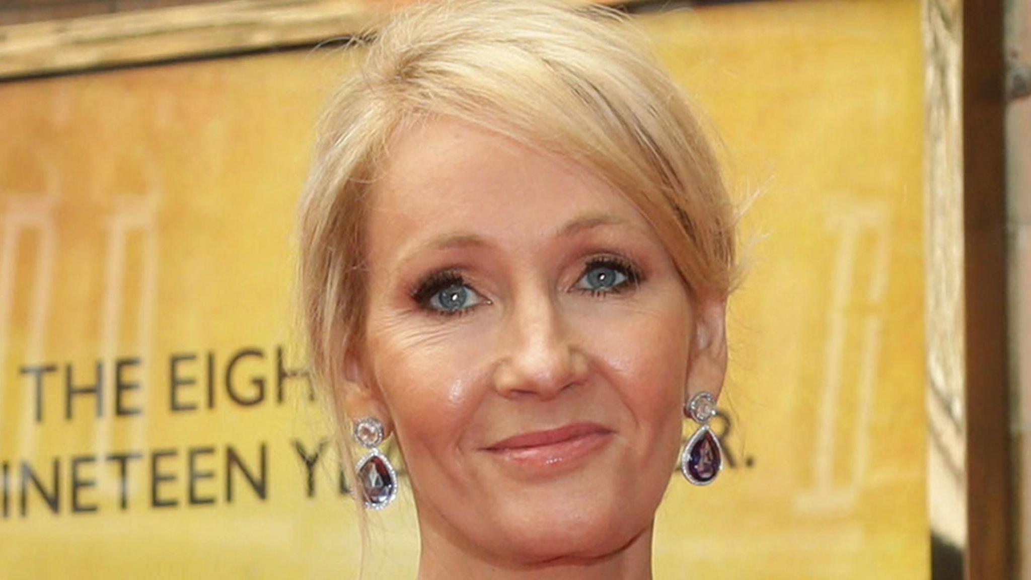 JK Rowling in close up in front of a gold-coloured backdrop. She is smiling, her hair is up and she is wearing lage ear rings.