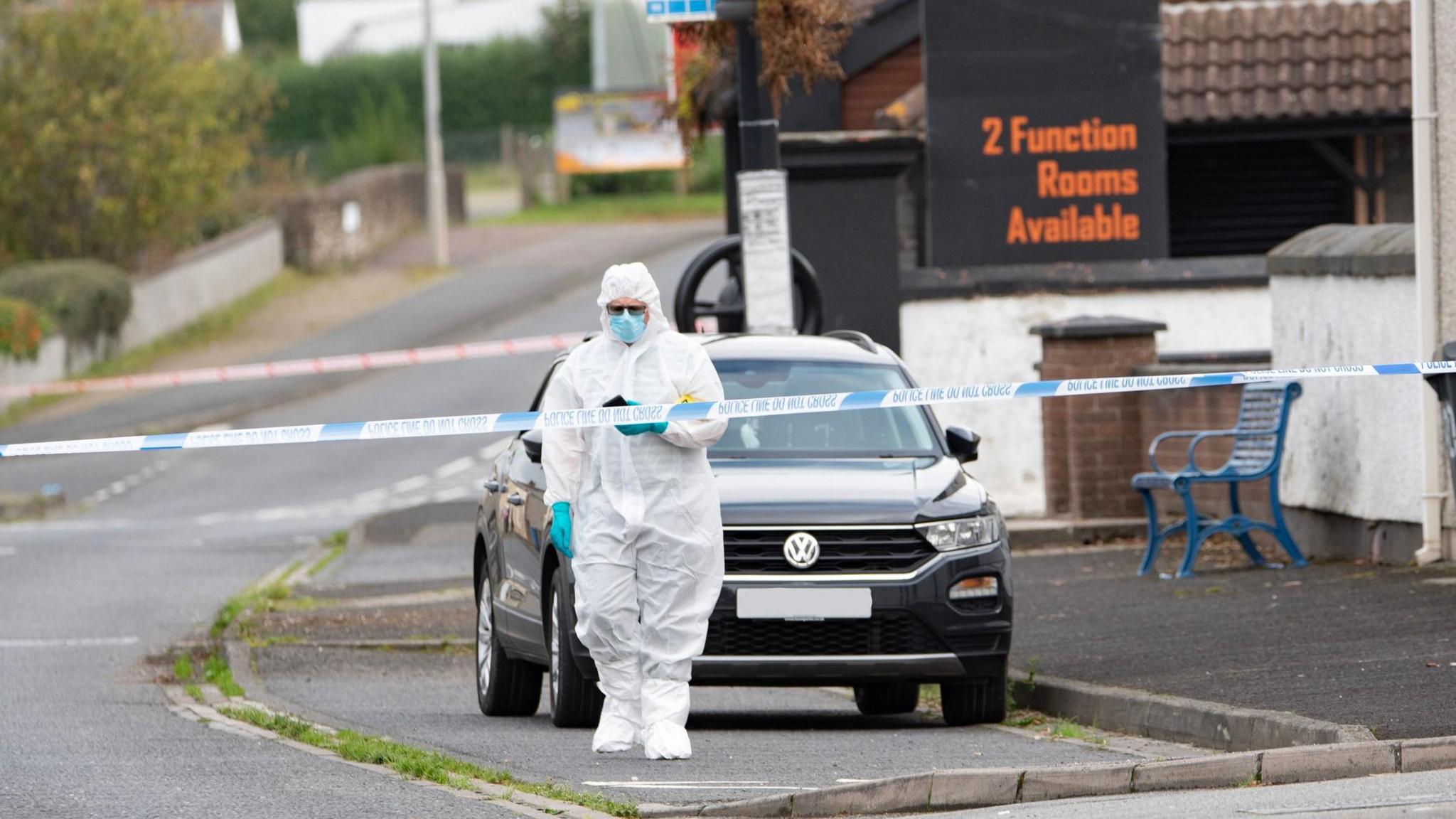 A police officer in a forensic suit stands in front of a black car, and beside a police tape. A number of houses are to the right, while a restaurant sign is in the background. A black wheely bin and park bench are also visible. 