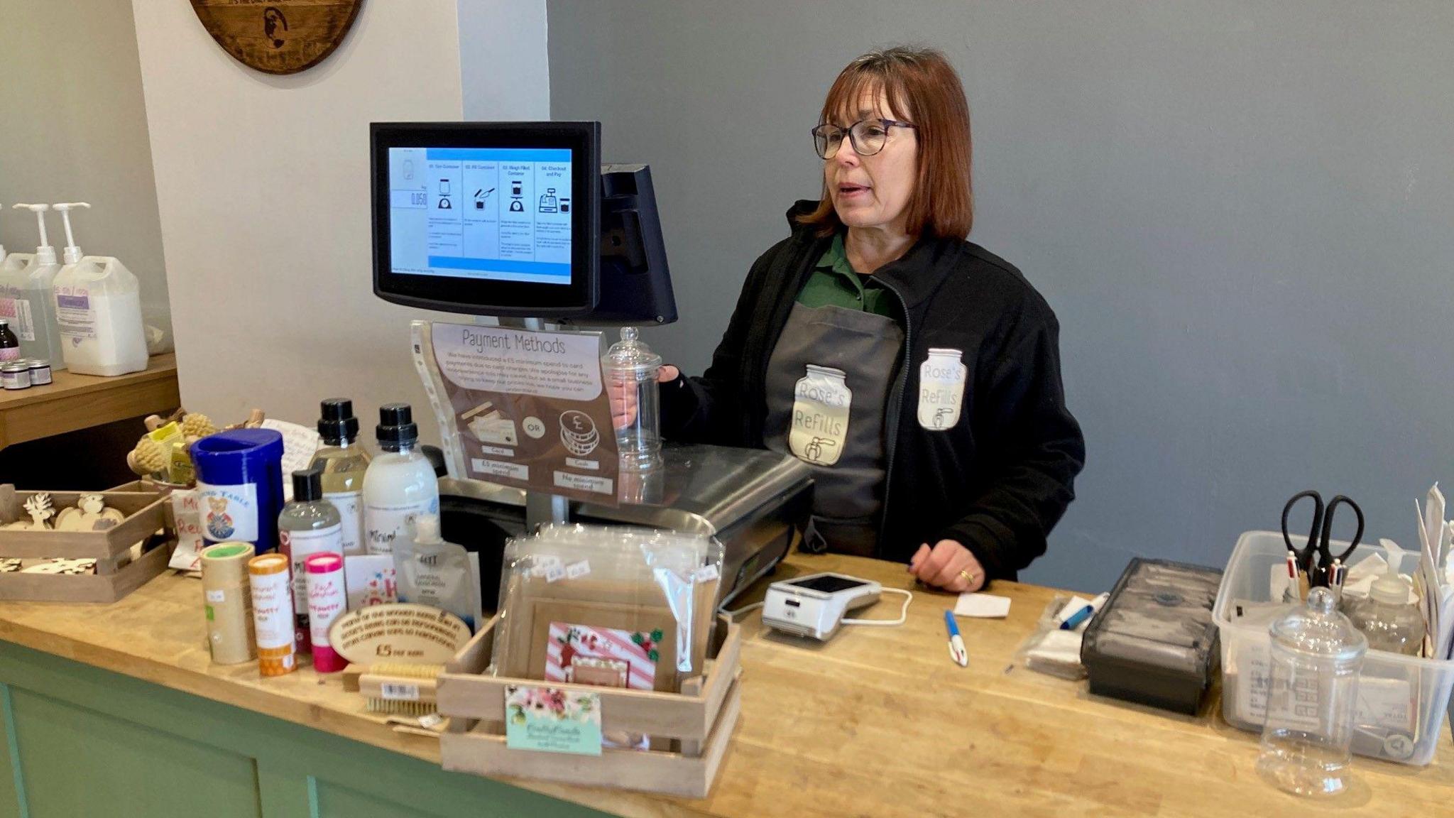 Rose King from Rose's Refills in her shop in Alsager.  She has dark red hair to her shoulders and wears round-rimmed glasses. She has a black fleece on over a grey apron which is over a green shirt. She is behind a wooden-topped counter with a range of shop-related items on it including a scales, a card reading machine, several bottles and a wooden basket with retail items in it.