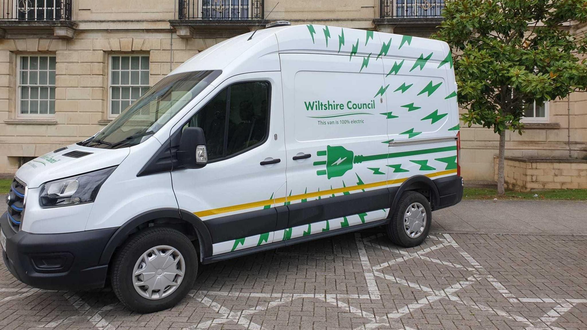 A white van in front of a building. The van has green electricity bolts over it and a large green plug, indicating it is an electric vehicle.