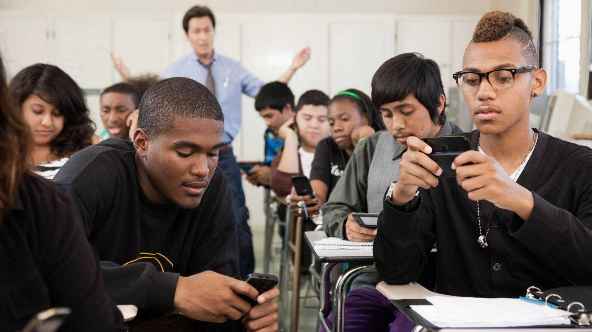 Students seated at desks in a classroom use cell phones as a teacher in the back of the class looks frustrated trying to get their attention