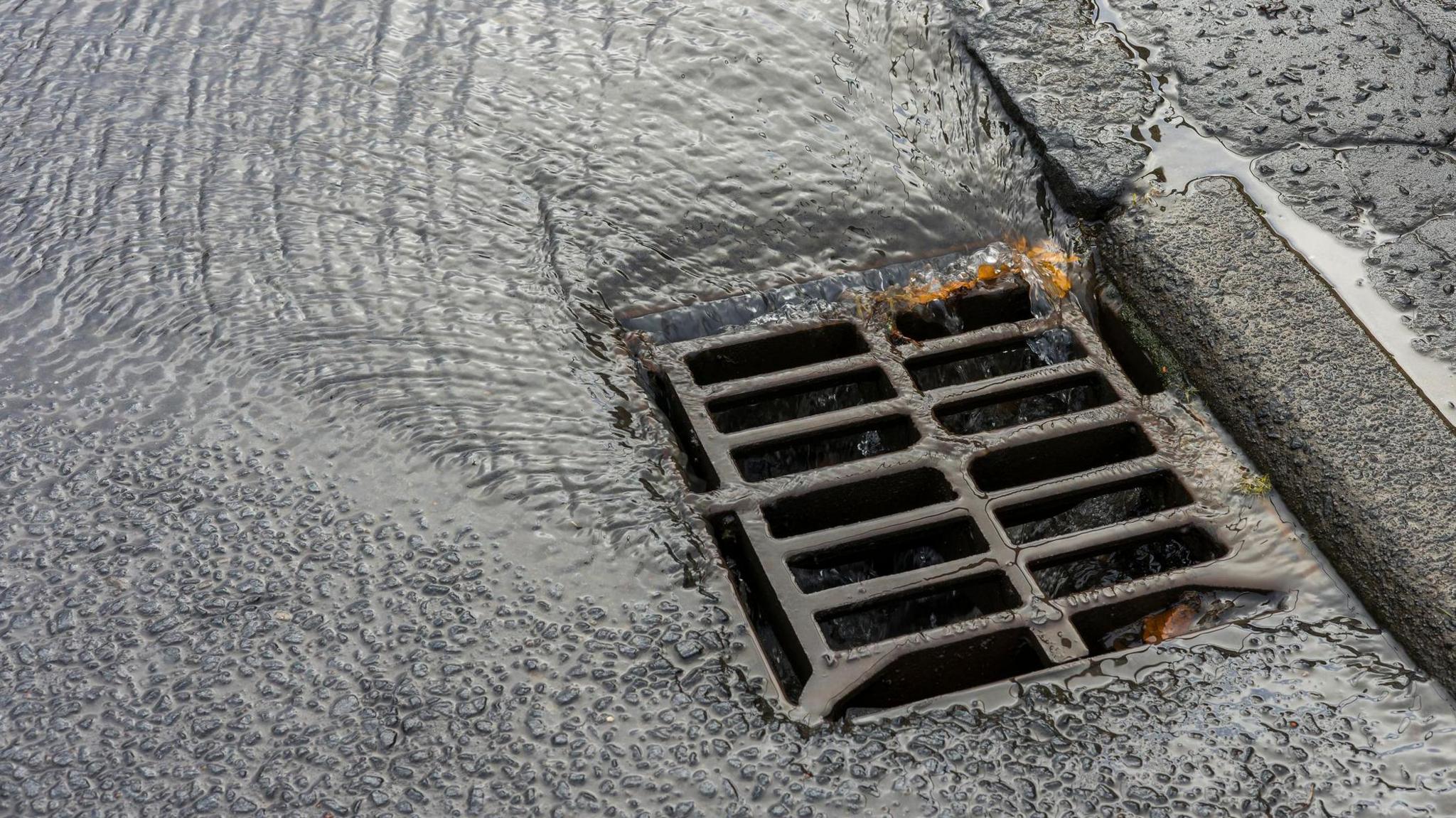 Close-up of a wet road gully with rainwater flowing into it. The surrounding road surface and kerb are slick with water after rainfall.