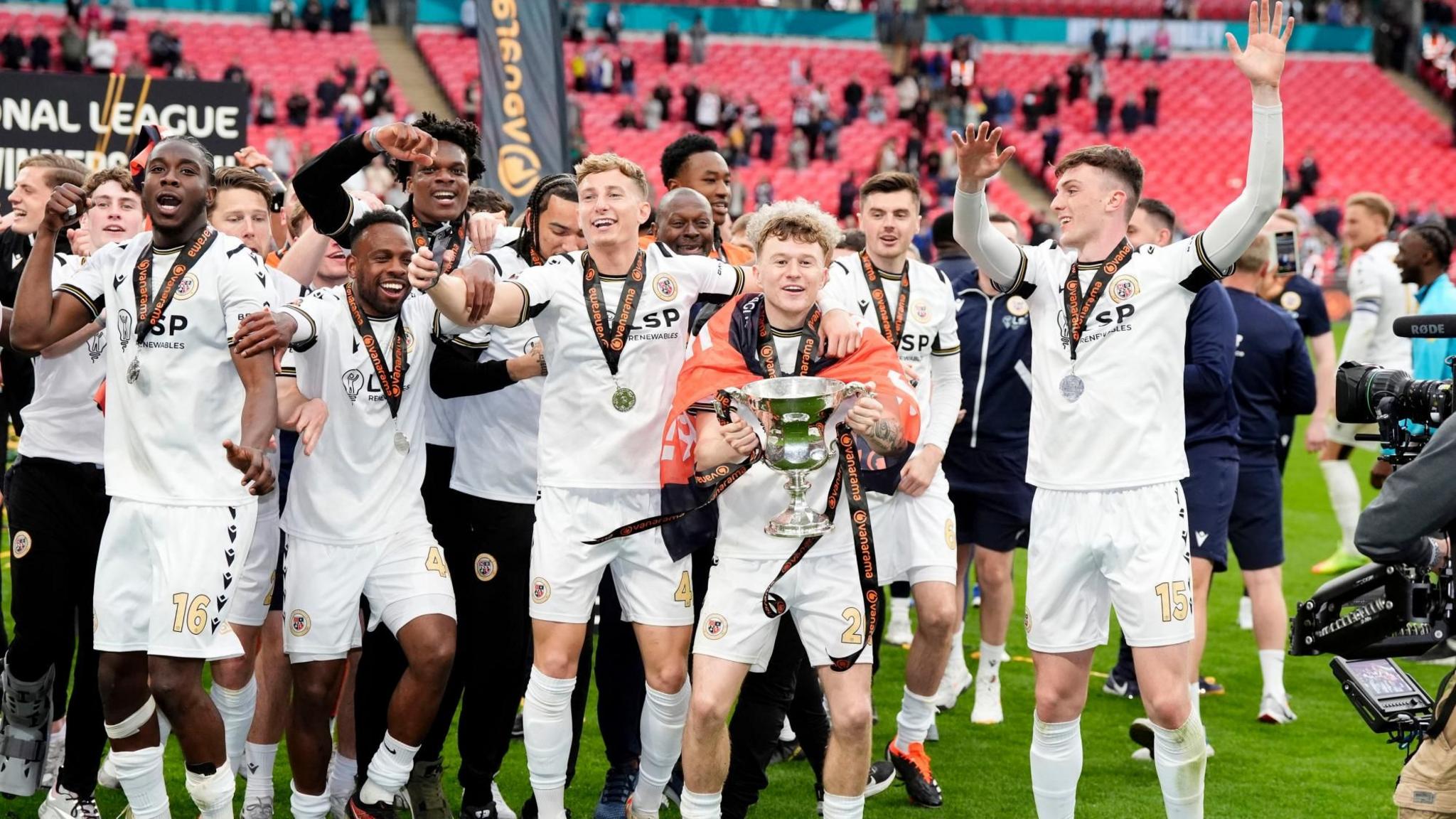 Bromley player in their all-white kit celebrate winning the National League play-off final at Wembley. One player holds the trophy while draped in an orange flag. A bank or empty red seats can be seen behind the players.