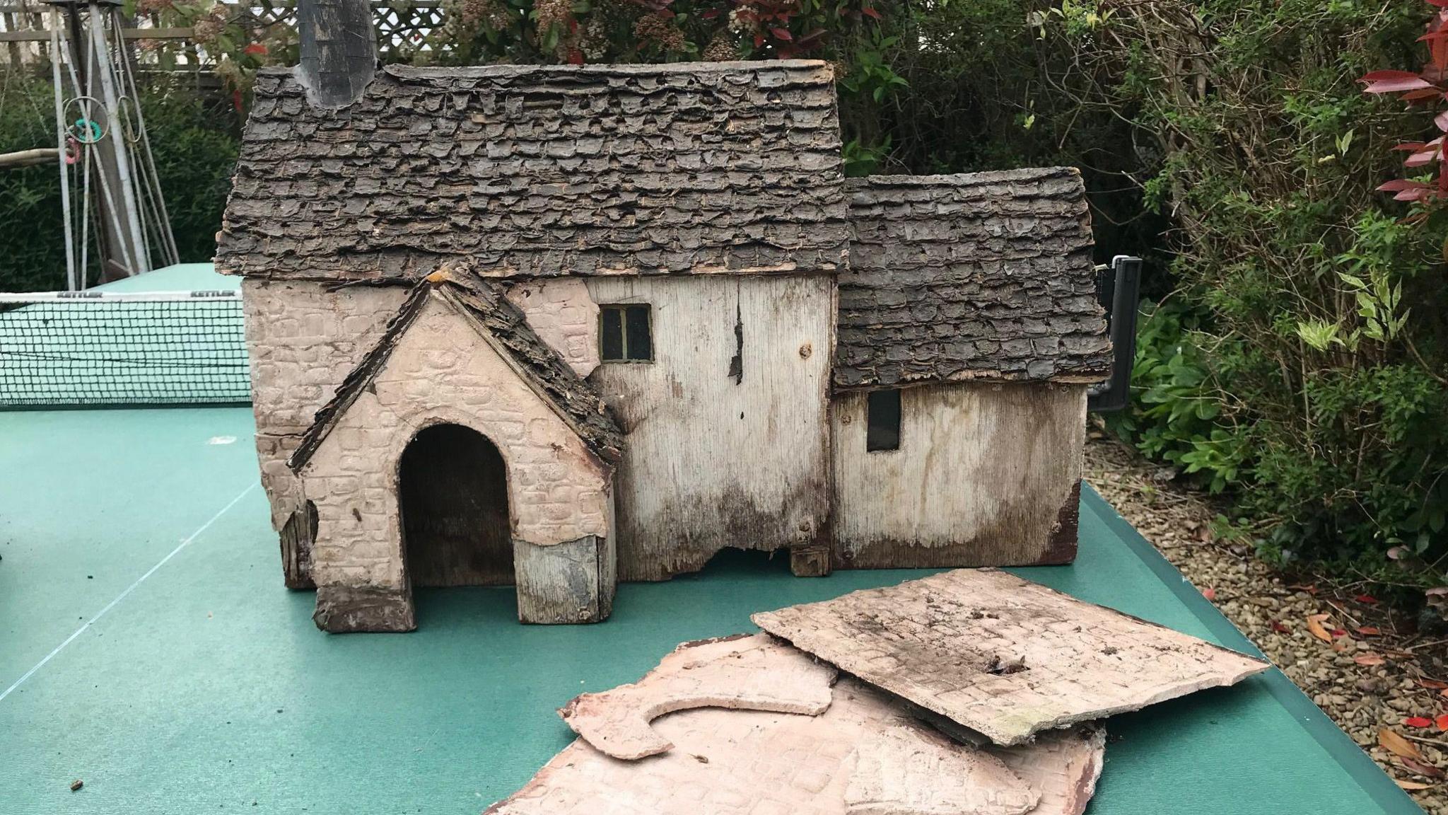 A miniature house is falling apart. Some of its walls have separated and it looks dilapidated. It's sitting on a green table tennis table.