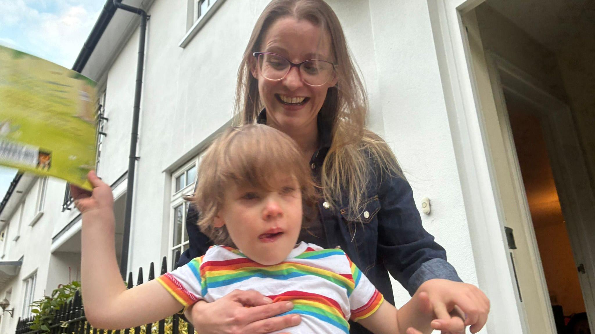 A young boy throwing a book. He is wearing a rainbow coloured striped shirt. He has wavy brown hair. His mother, who is holding him, is wearing glasses and a dark blue denim top.