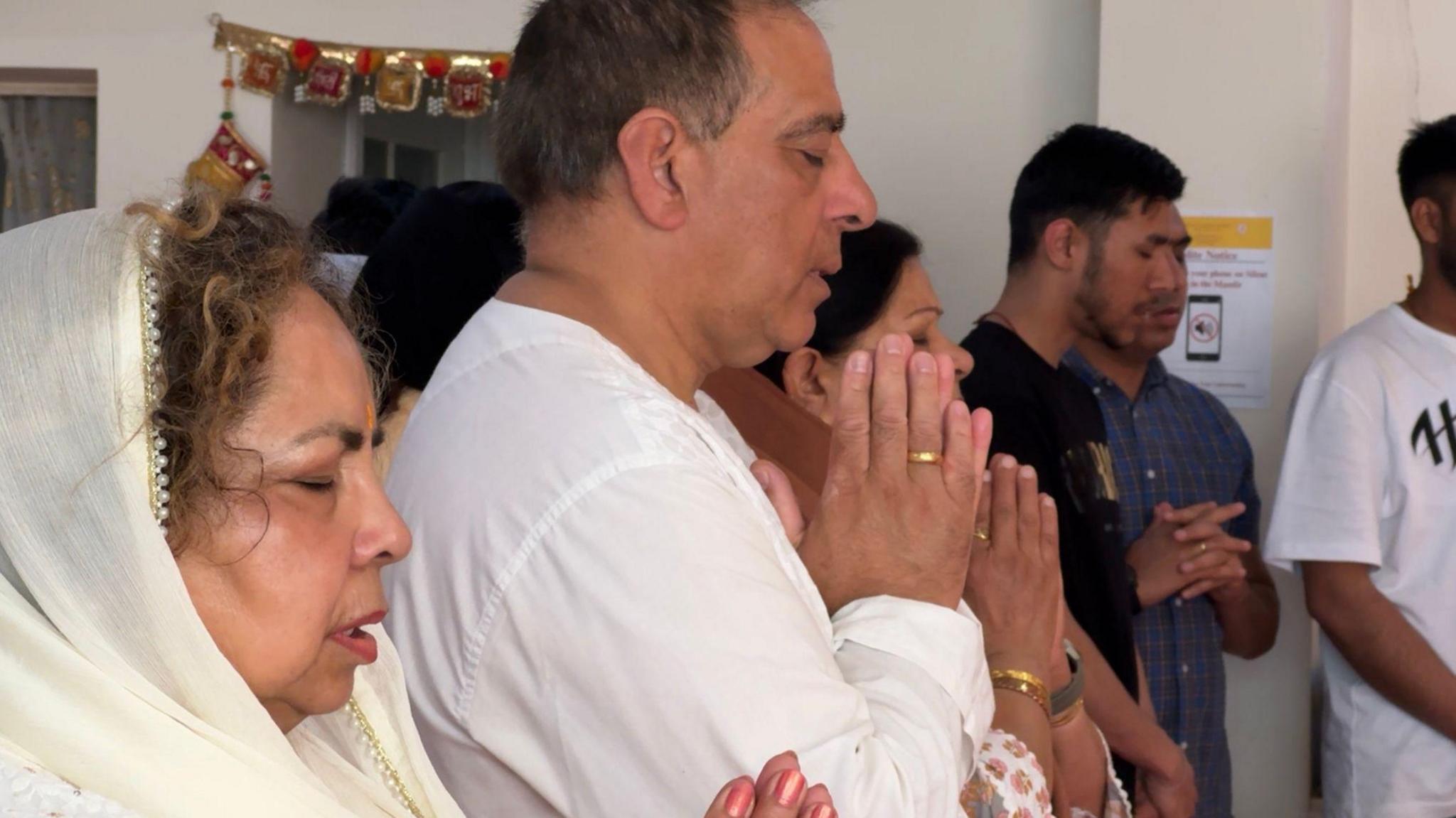 A group of people praying. They are mainly wearing white and have their hands pressed together in front of them in a prayer symbol.